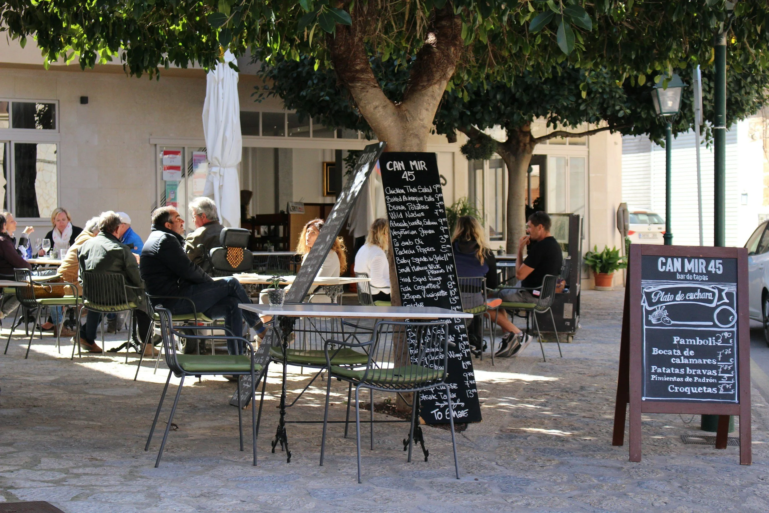 Outdoor cafe with several people sitting at tables under trees, with handwritten chalkboard menus displaying Spanish food and drink options.