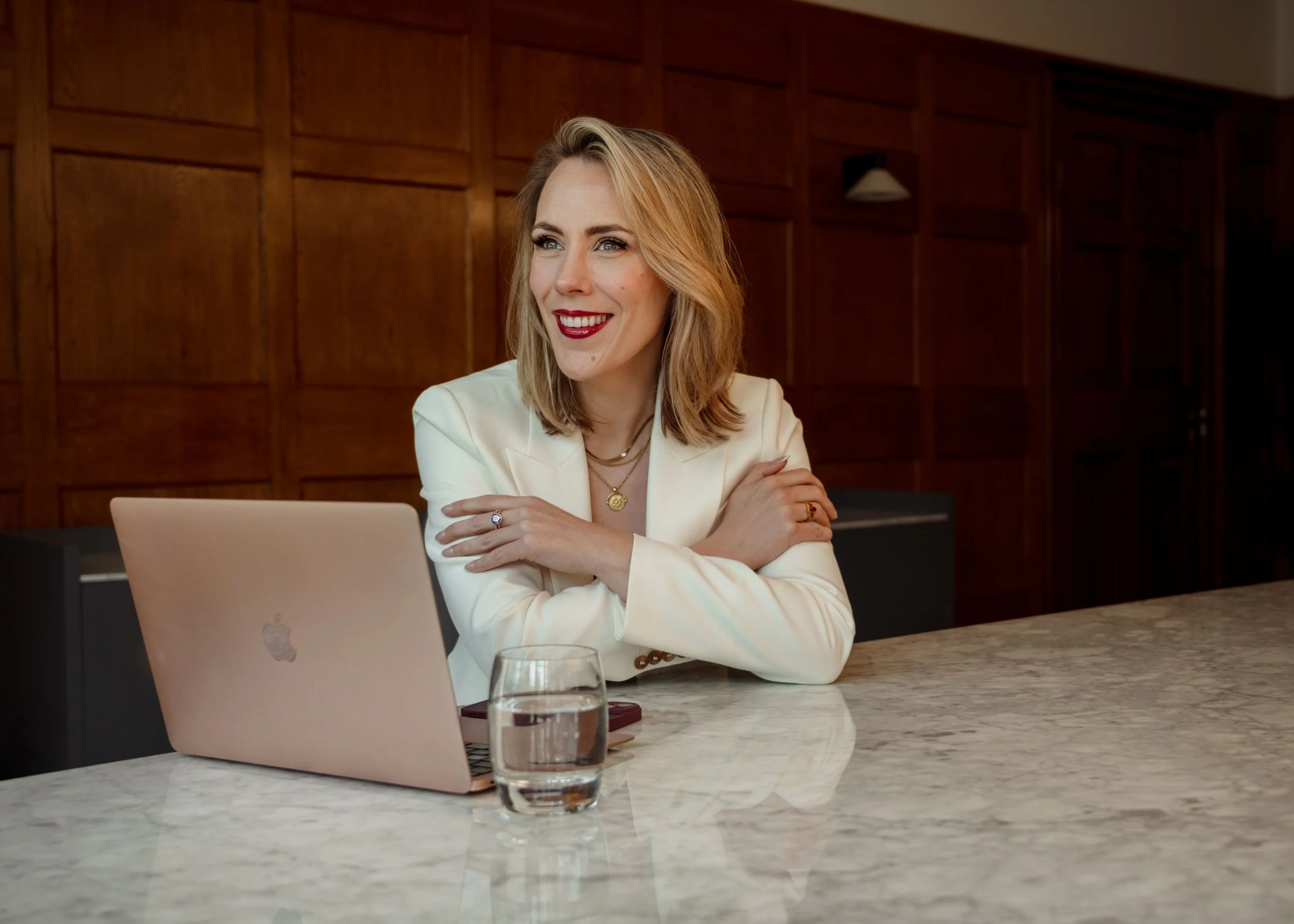 Alex Thornton - Founder, Coach and Leadership Development Specialist. A woman with blond hair wearing a cream suit, sitting at a marble table with a laptop, glass of water, and smartphone, smiling and looking to her right, in a wood-paneled room.