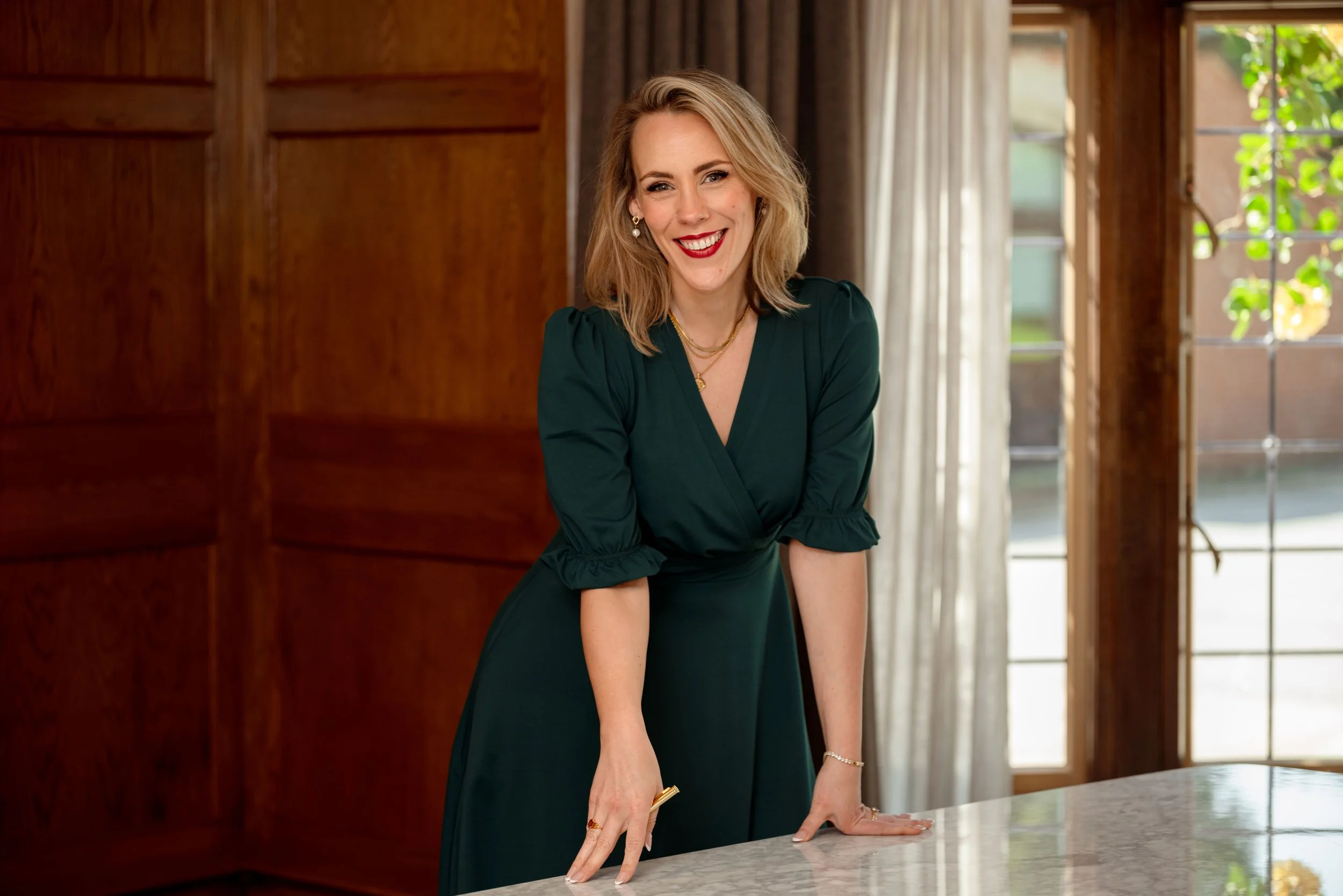 Alex Thornton - Founder, Coach and Leadership Development Specialist. A smiling woman with blonde hair and red lipstick leaning on a white marble countertop in a warmly lit room with wooden-paneled walls and large windows.