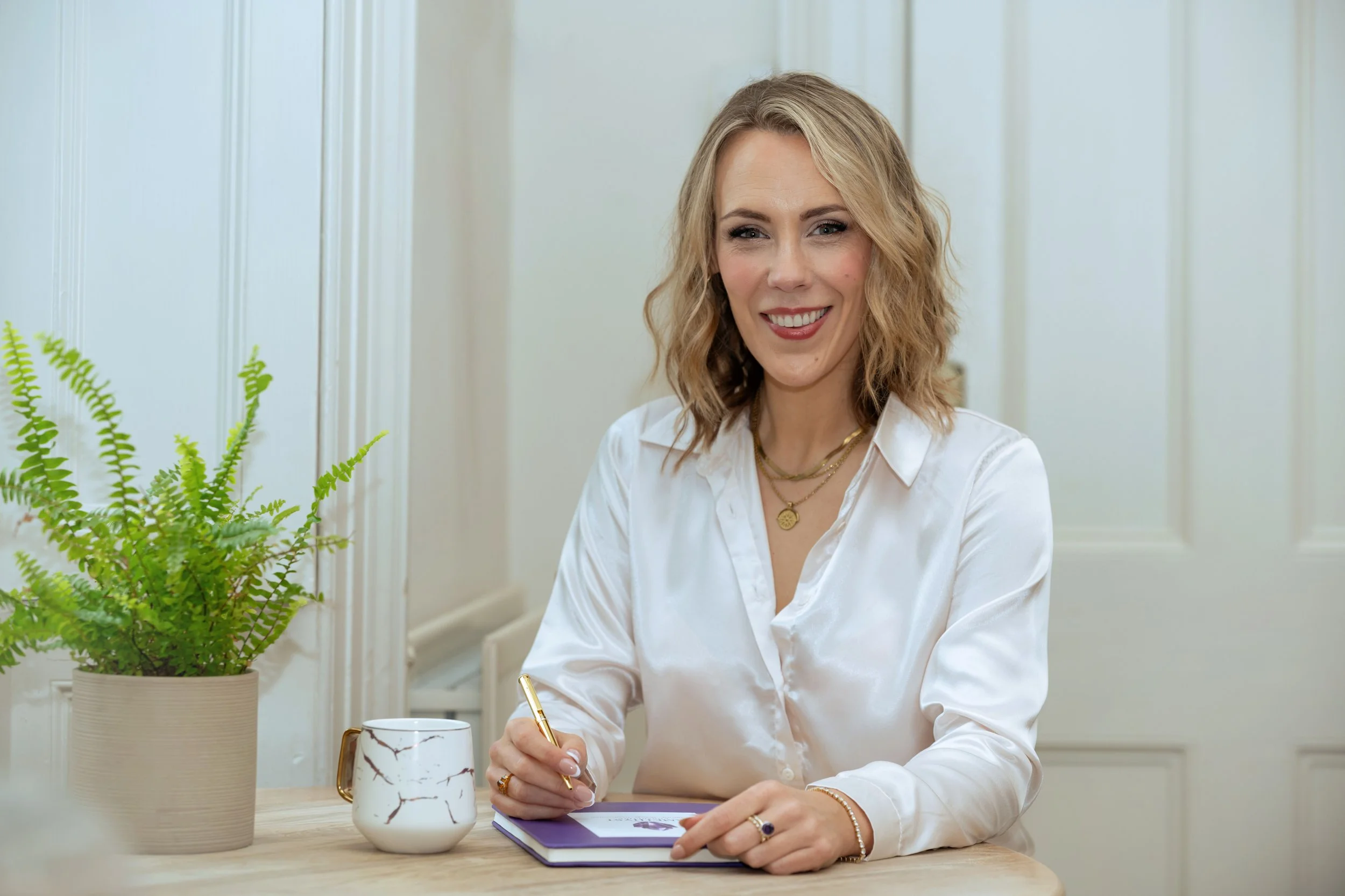 Alex Thornton - Founder, Coach and Leadership Development Specialist. A woman with blonde, wavy hair wearing a white blouse, sitting at a table, smiling, with a plant and a mug nearby, in a bright room.