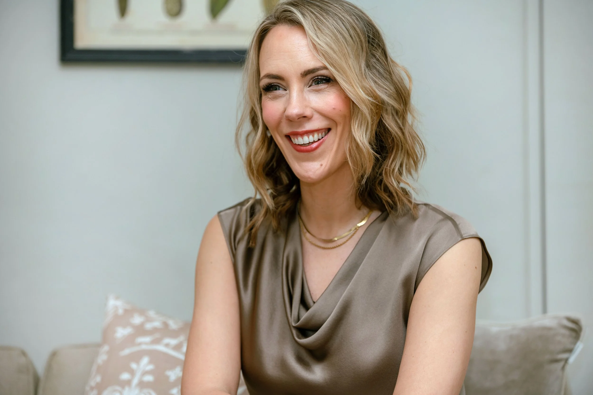 Alex Thornton - Founder, Coach and Leadership Development Specialist. A woman smiling with blonde wavy hair, wearing a taupe satin top and gold jewelry, sitting indoors with a framed art piece on the light-colored wall behind her.