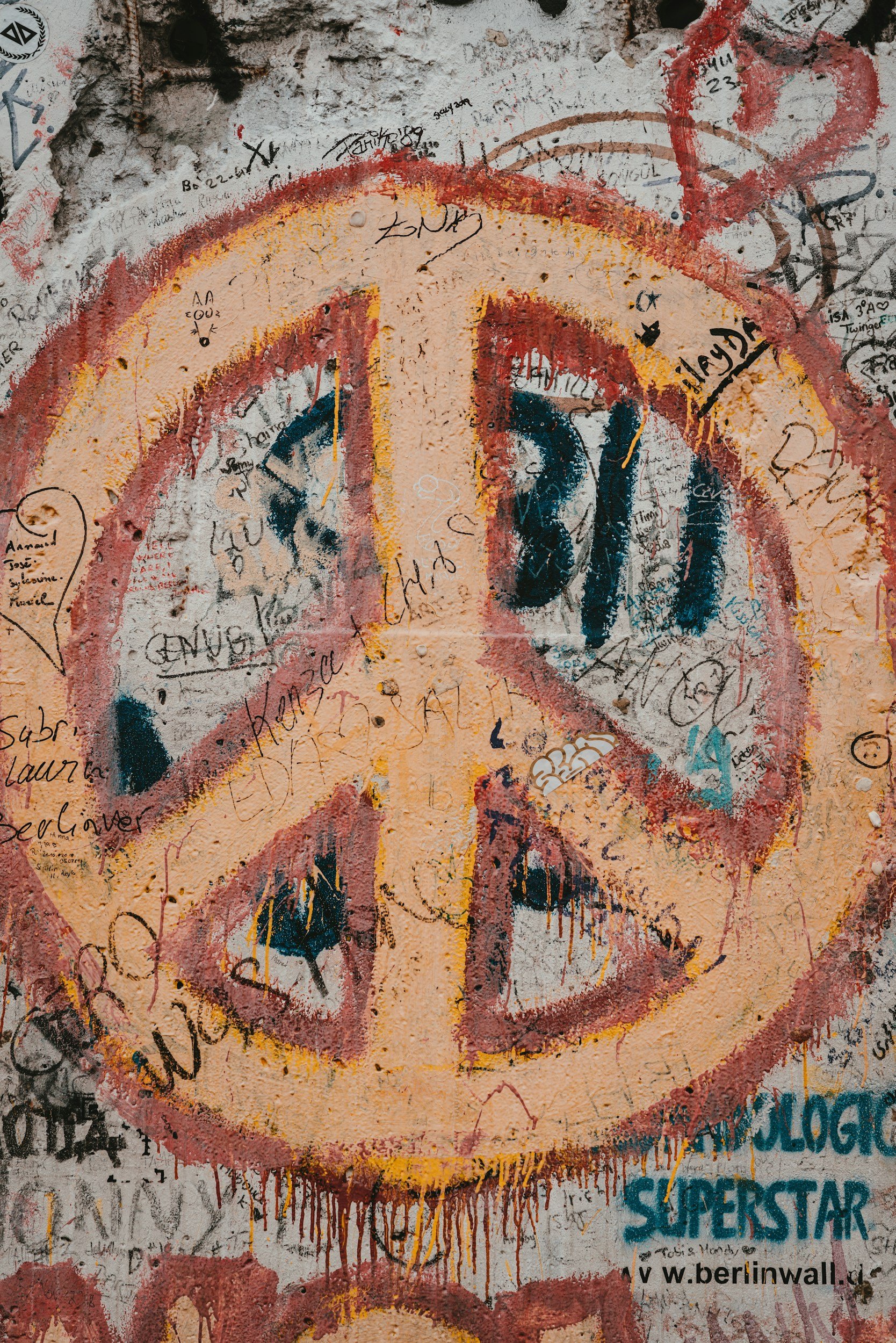 Graffiti art on a wall depicting a large, stylized smiley face. The face is outlined in red and yellow, with black eyes and a wide, black, smiling mouth. The wall is covered with various handwritten notes, signatures, and graffiti tags in different colors, overlapping each other.
