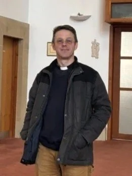 A man standing indoors, wearing glasses, a black jacket, and khaki pants, smiling at the camera.