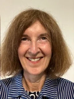A woman with light skin, shoulder-length curly brown hair, smiling and wearing a blue and white striped shirt.