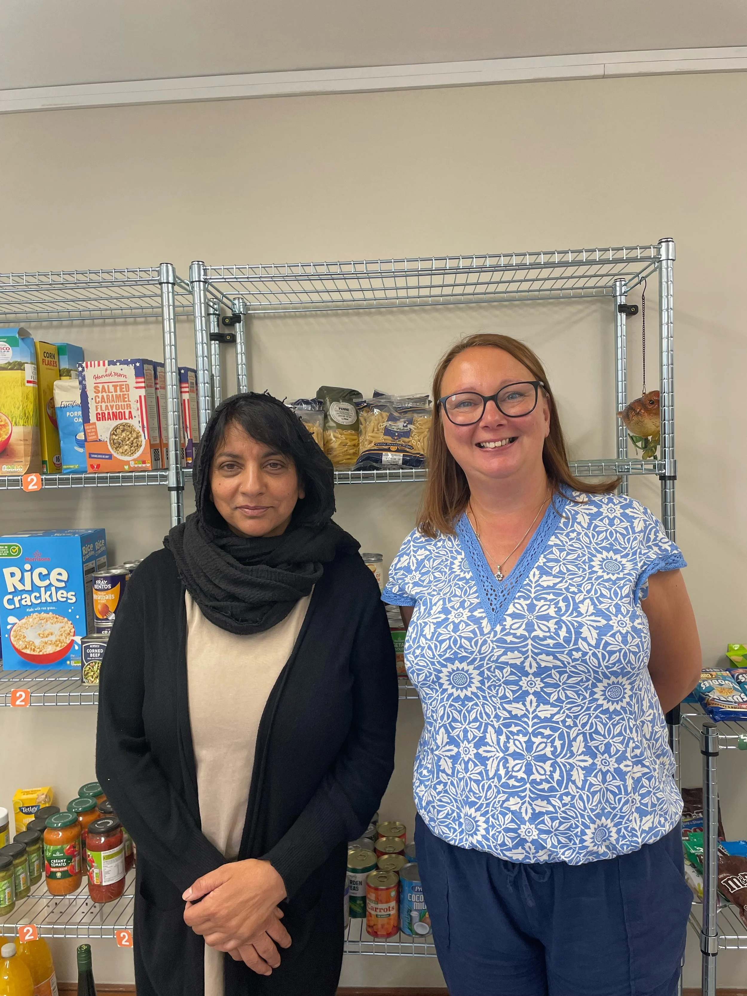 Two women standing in front of a metal grocery shelf with various canned and boxed foods. One woman is wearing a black hoodie and scarf, the other is wearing glasses and a blue and white patterned shirt.