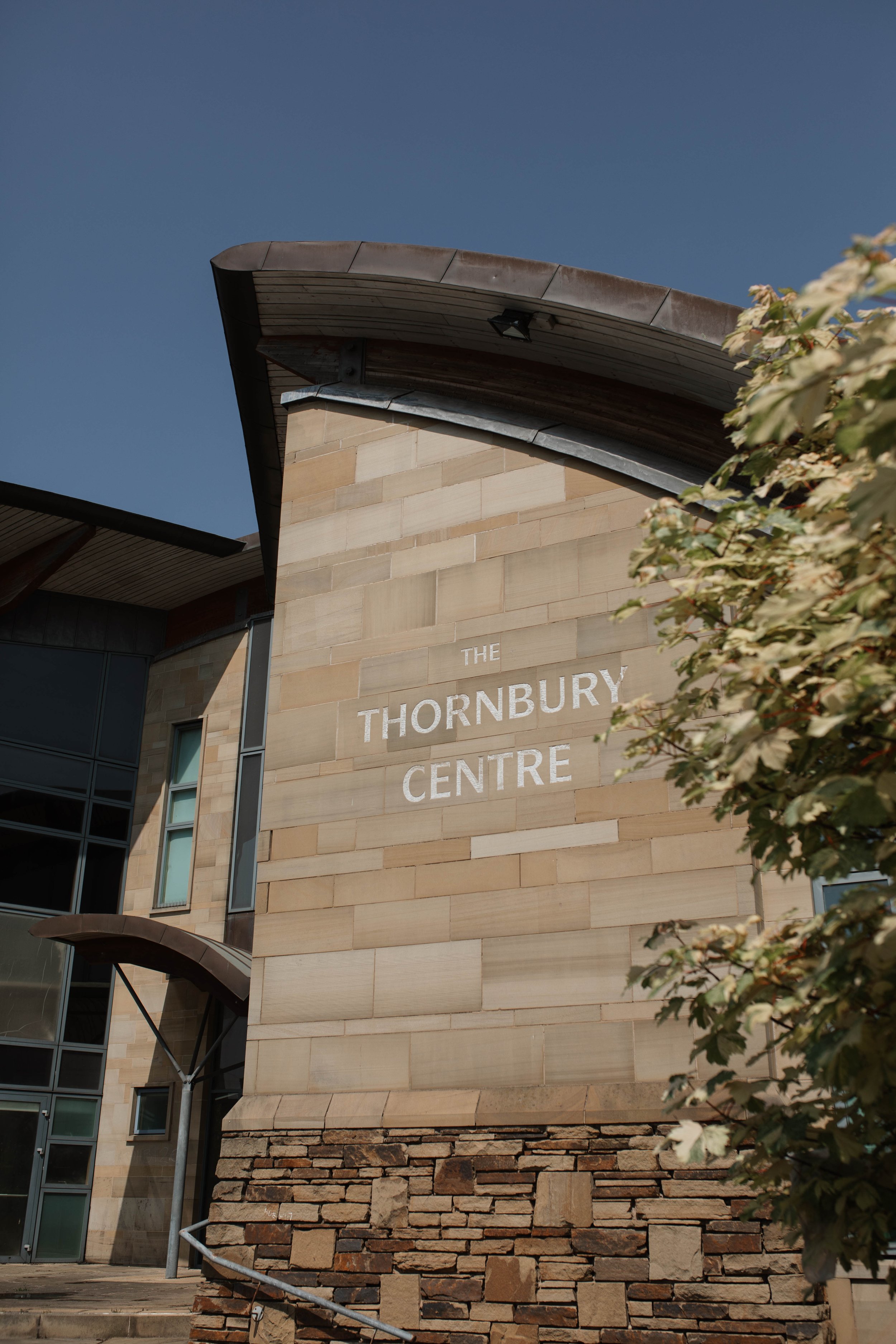 Close-up view of the exterior of the Thornbury Centre building with beige brick and stone wall, text reading 'THE THORNBURY CENTRE', overcast sky, and part of tree with leaves in the right foreground.