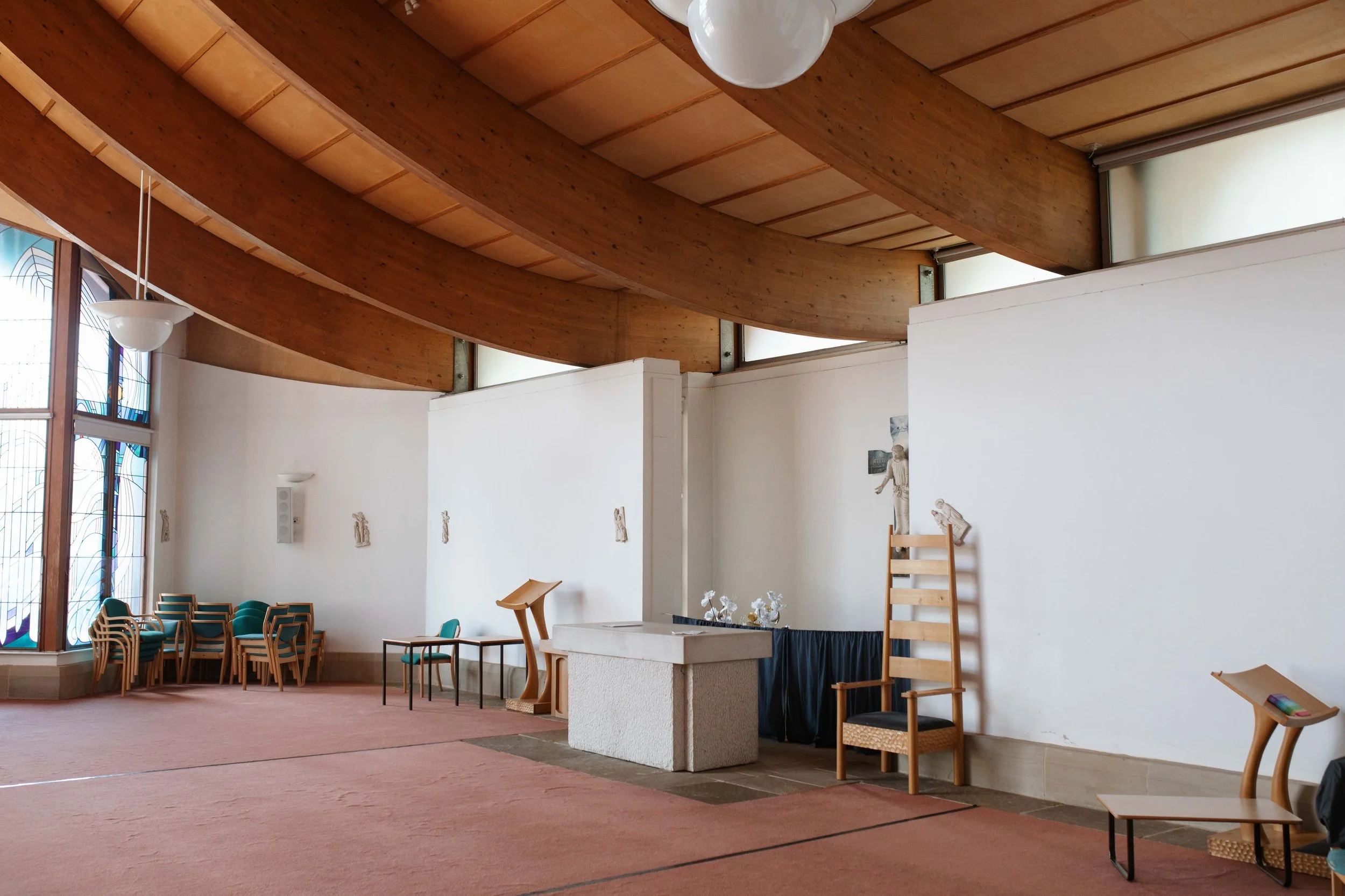 Interior of a church or chapel featuring wooden ceiling beams, stained glass windows, and simple furniture including chairs and small tables.