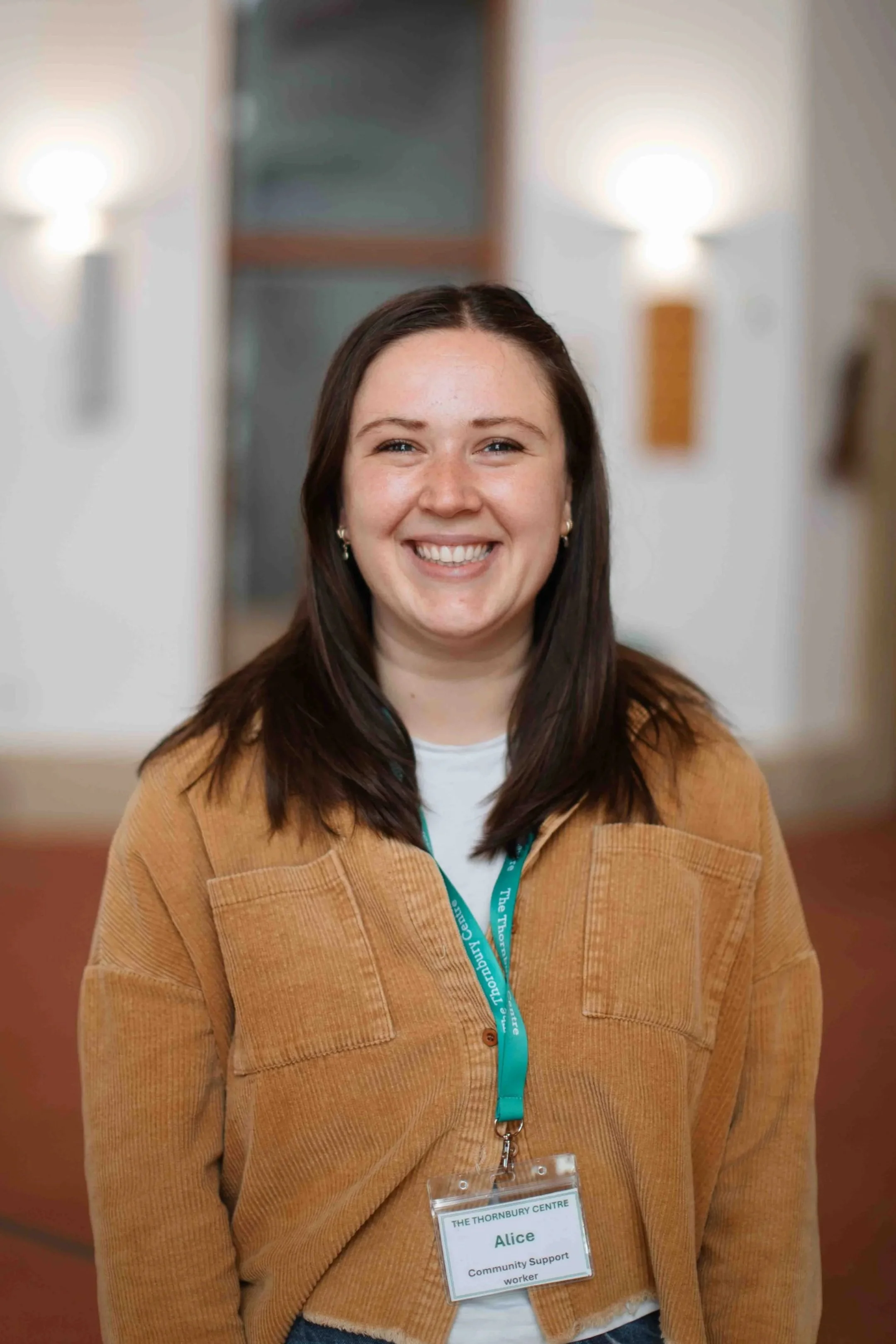A smiling woman with brown hair, wearing a tan jacket and a name badge, standing in a room with blurred background lighting.