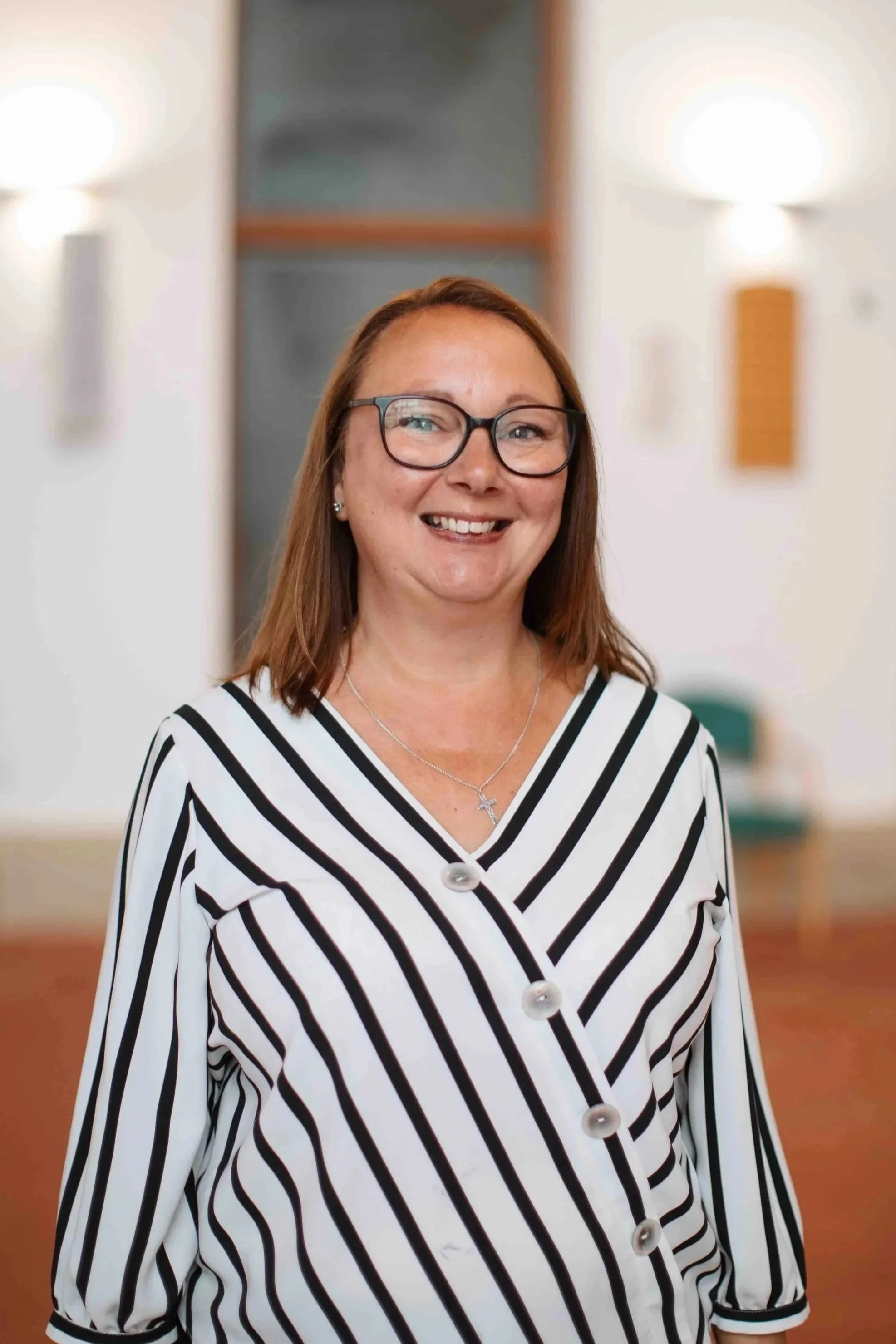 A smiling woman with red hair and glasses, wearing a black and white striped blouse, standing indoors with blurred lights and background.