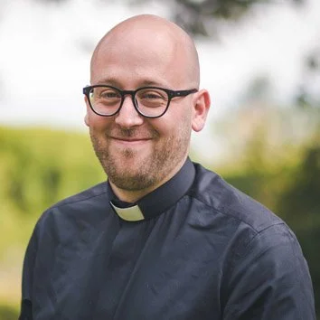 A smiling man with glasses and a clerical collar, outdoors with a blurred green background.