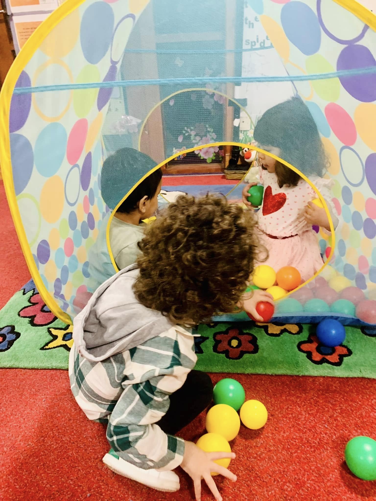 Child playing with colorful plastic balls inside a small play tent with a polka dot pattern, on a carpeted floor.
