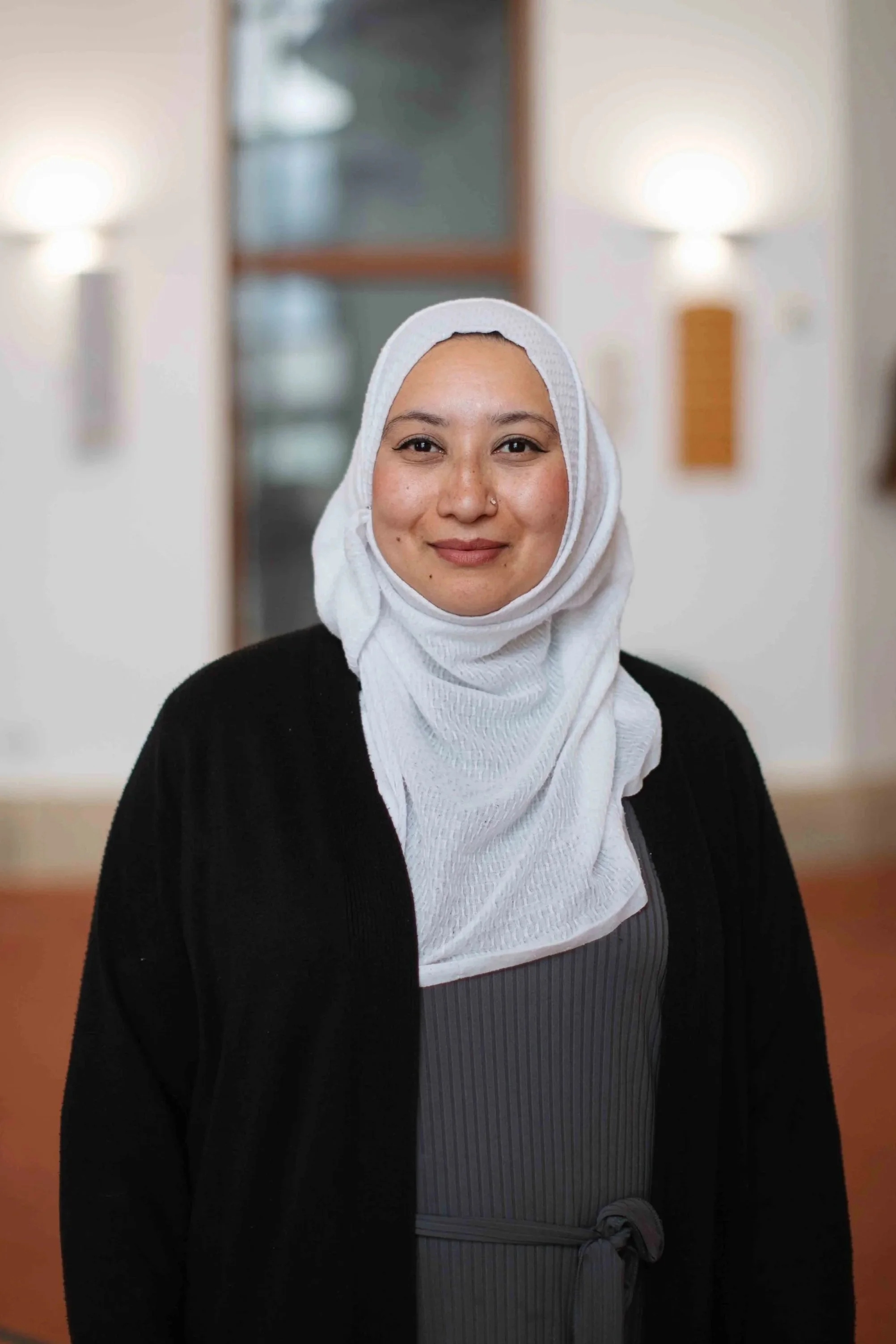 A woman with a light brown complexion, wearing a white hijab, a gray dress, and a black jacket, standing in a room with blurred background featuring two wall sconces.