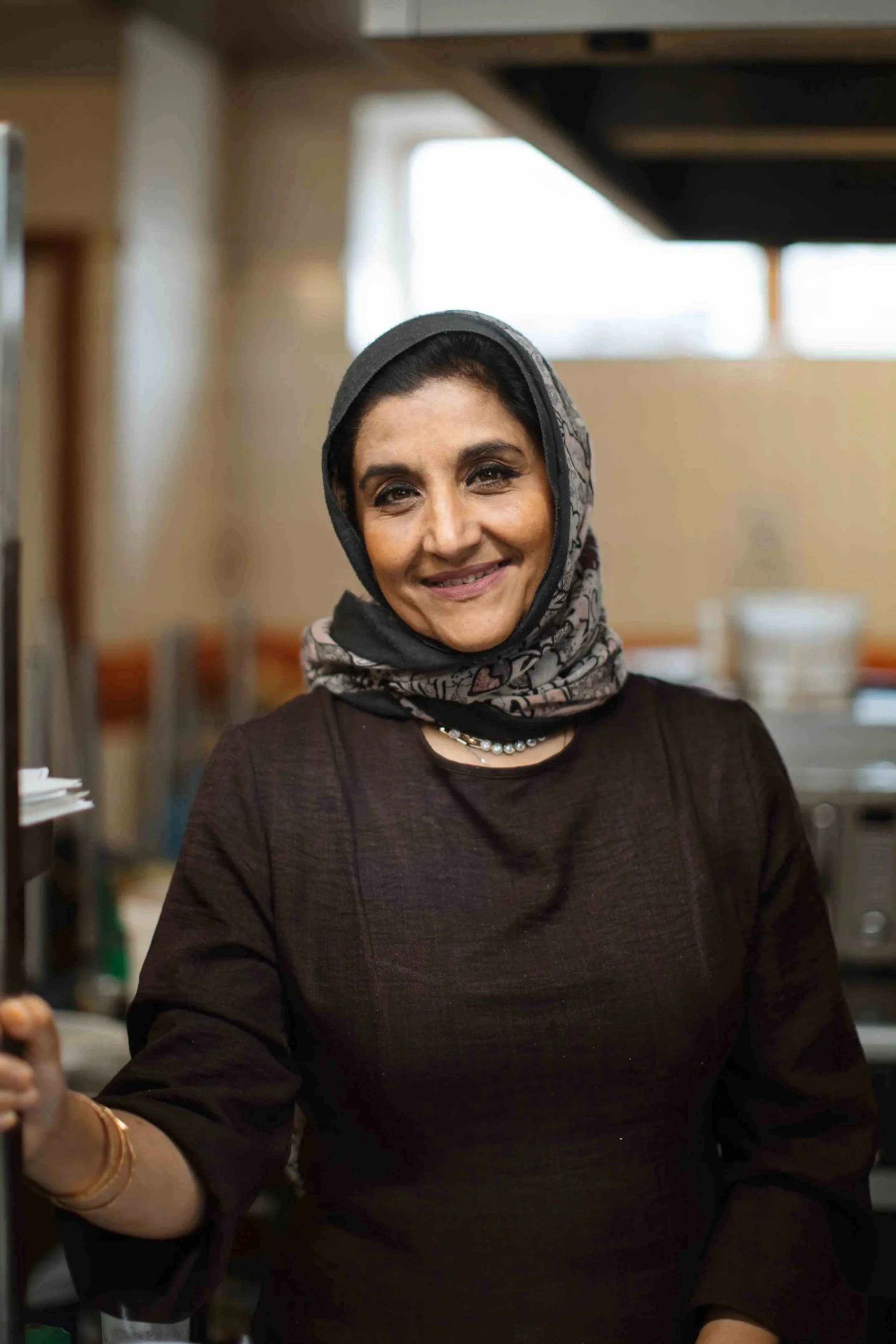 A woman wearing a dark sweater and a patterned gray headscarf, smiling in a professional kitchen.