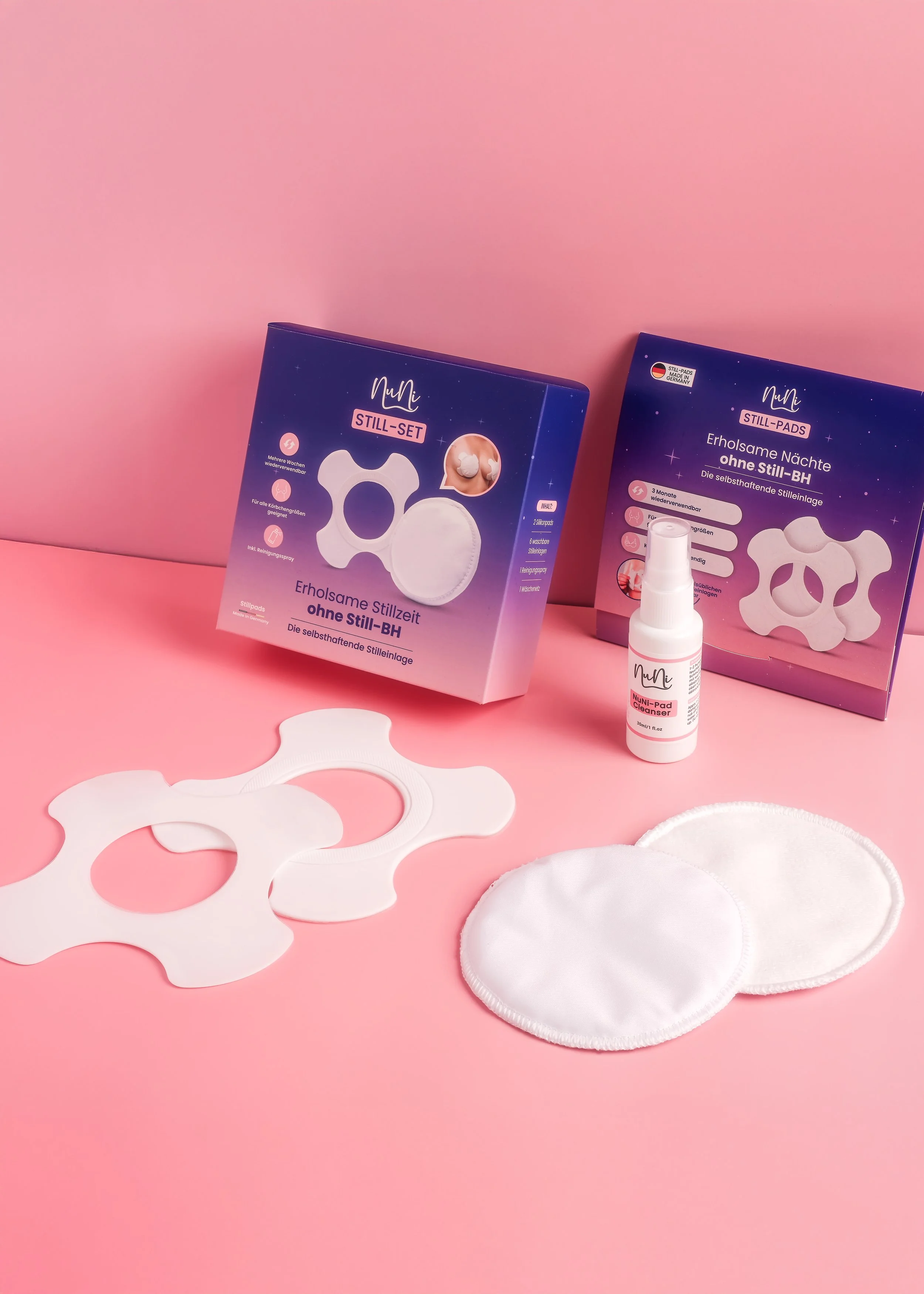 Pink background with boxes of postpartum still pads, white disposable breast pads, and a small spray bottle labeled NuNi-Pad Cleaner.