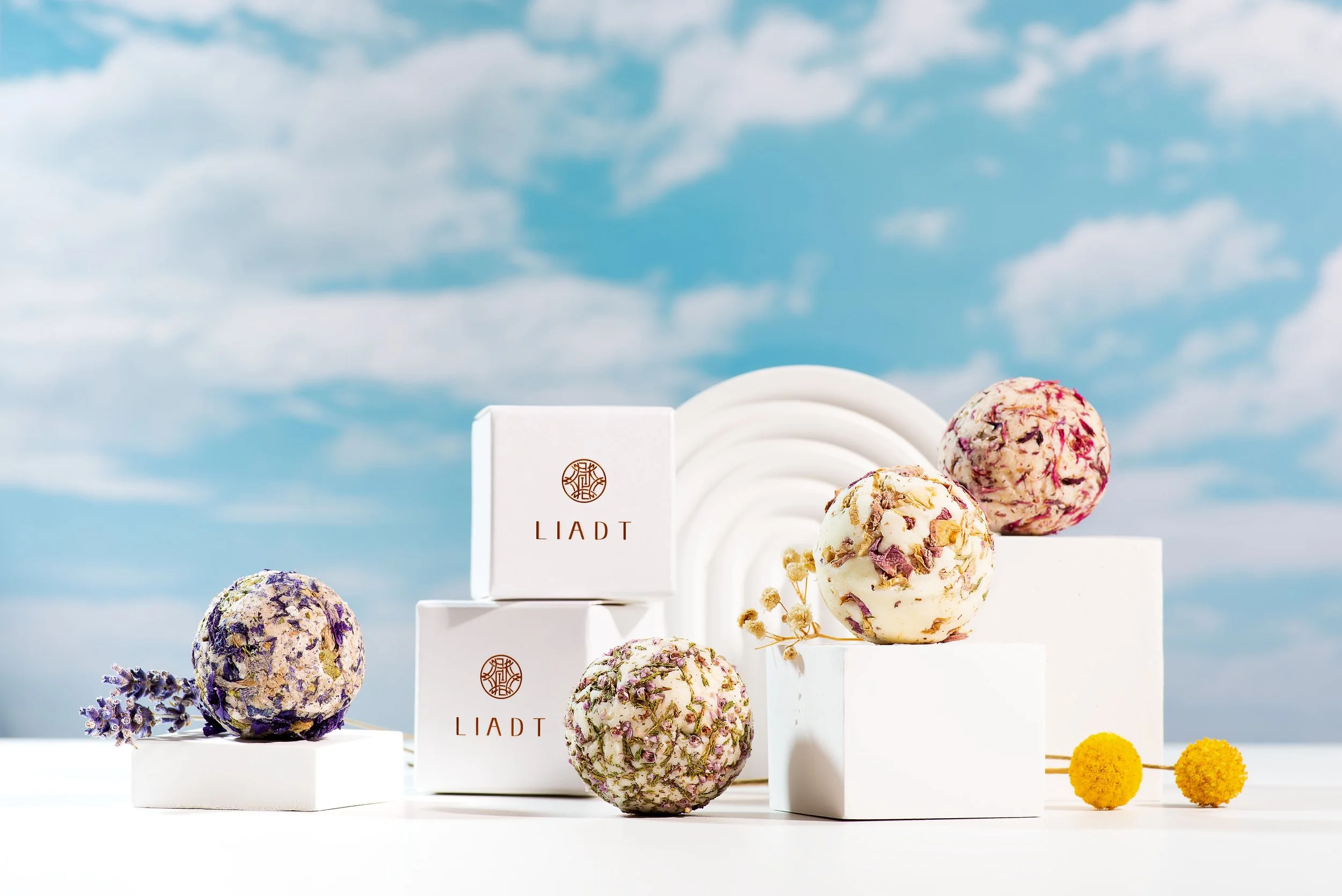 Six colorful, flower-embellished ice cream scoops on white display blocks with lavender and yellow decorative elements against a blue sky with clouds.