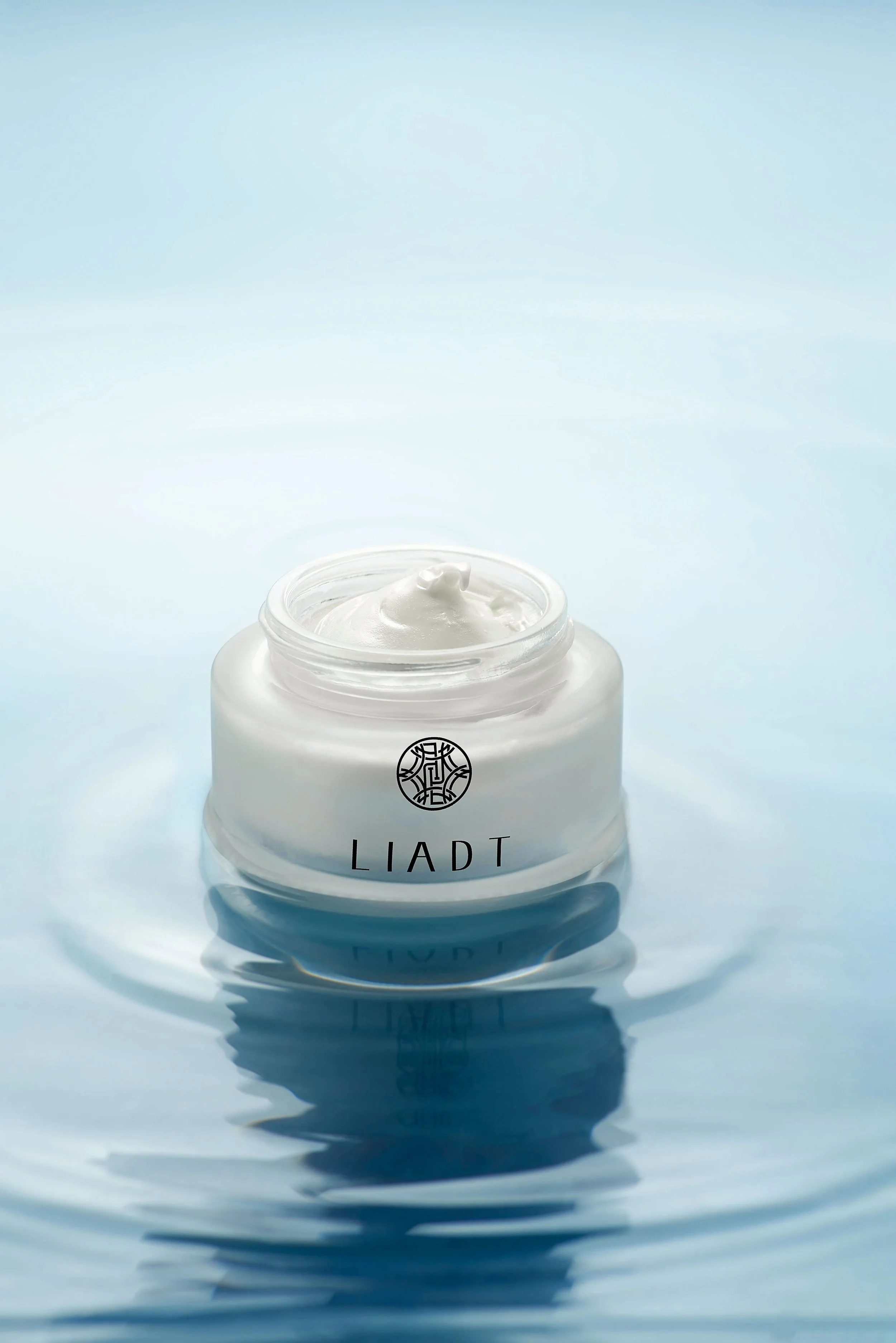 A jar of LIADT skincare cream with a swirl of cream on top, reflected on a surface with a light blue background.