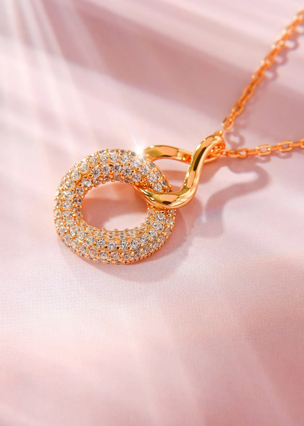 Gold necklace with a circular pendant encrusted with small diamonds, resting on pink satin fabric.