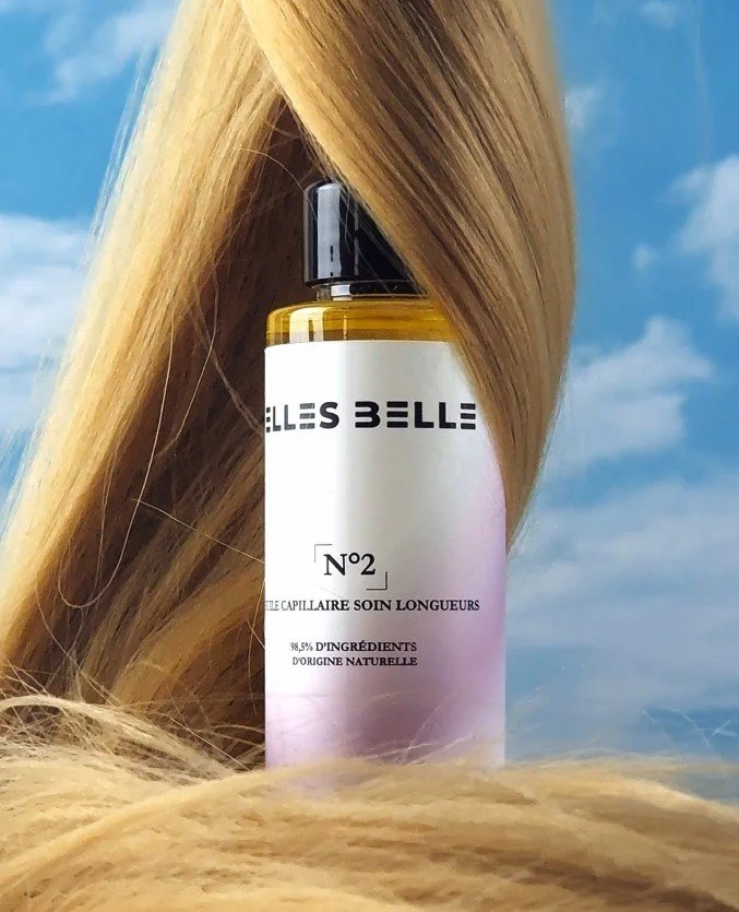 A bottle of Belle Belle hair product placed outdoors with blonde hair draped around it and blue sky with clouds in the background.
