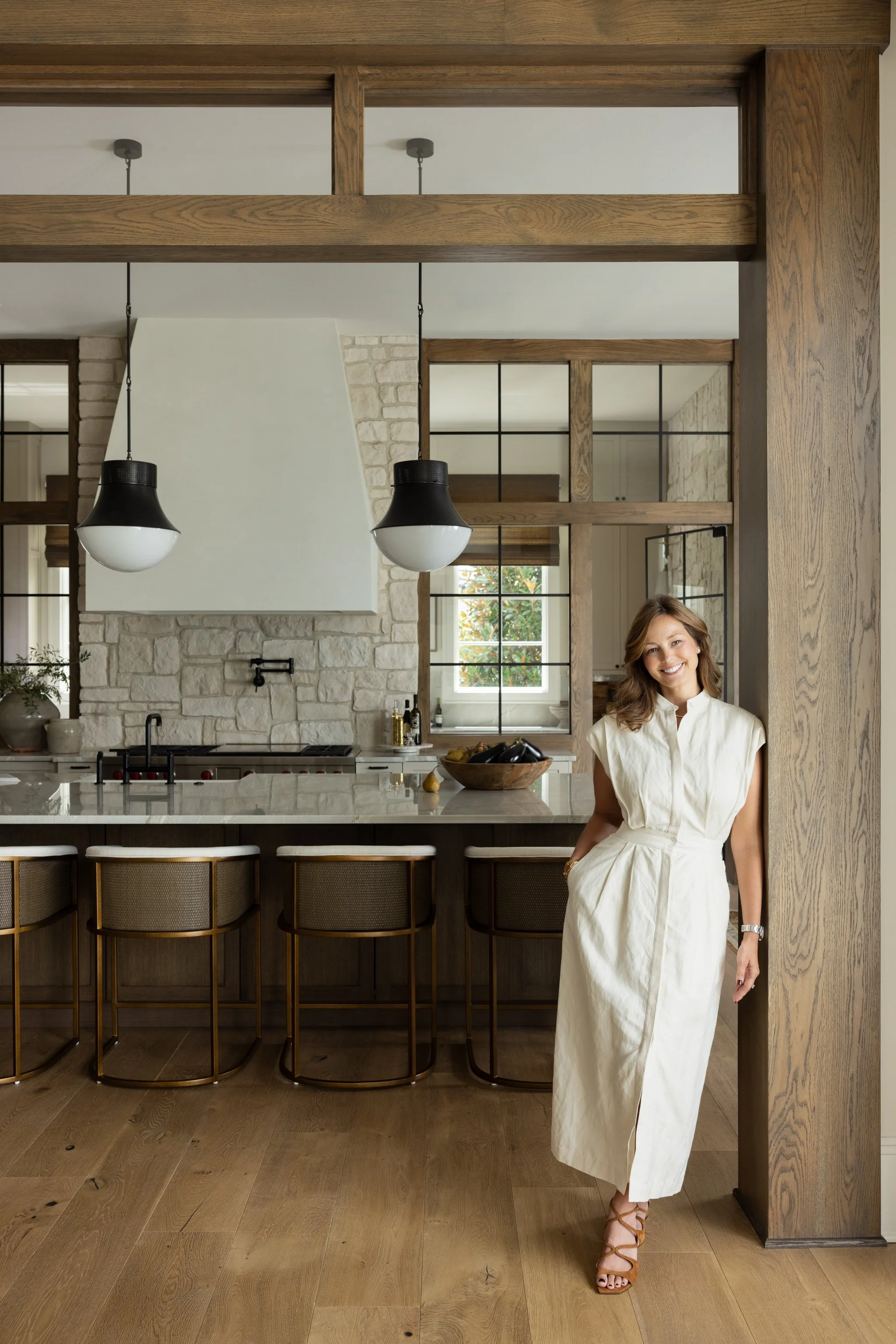 A woman in a white dress standing in a modern kitchen with wooden accents, stone wall, black and white pendant lights, and four bar stools.