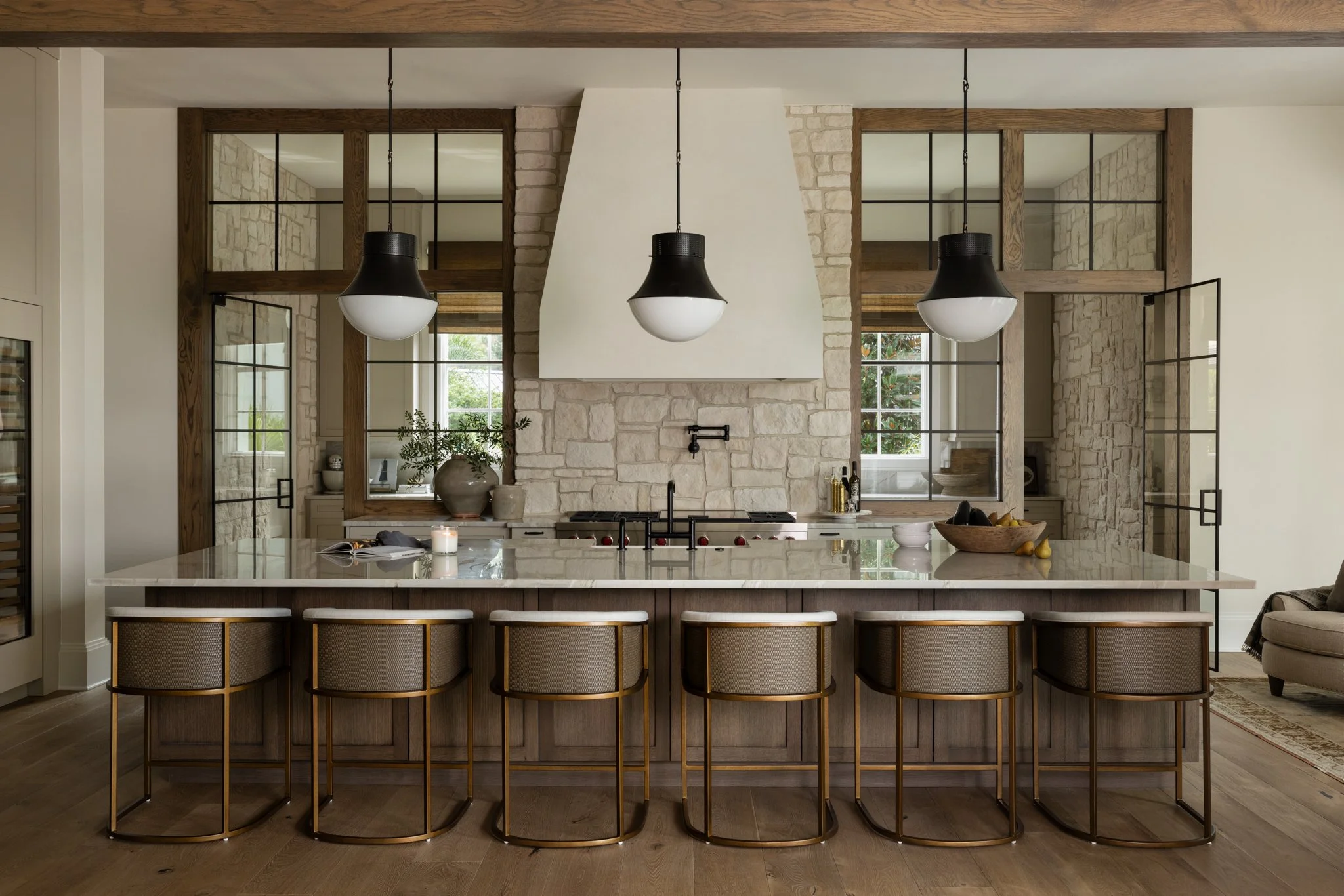 Modern kitchen with a large island, high-back chairs, pendant lighting, stone wall, and windows with wooden frames.