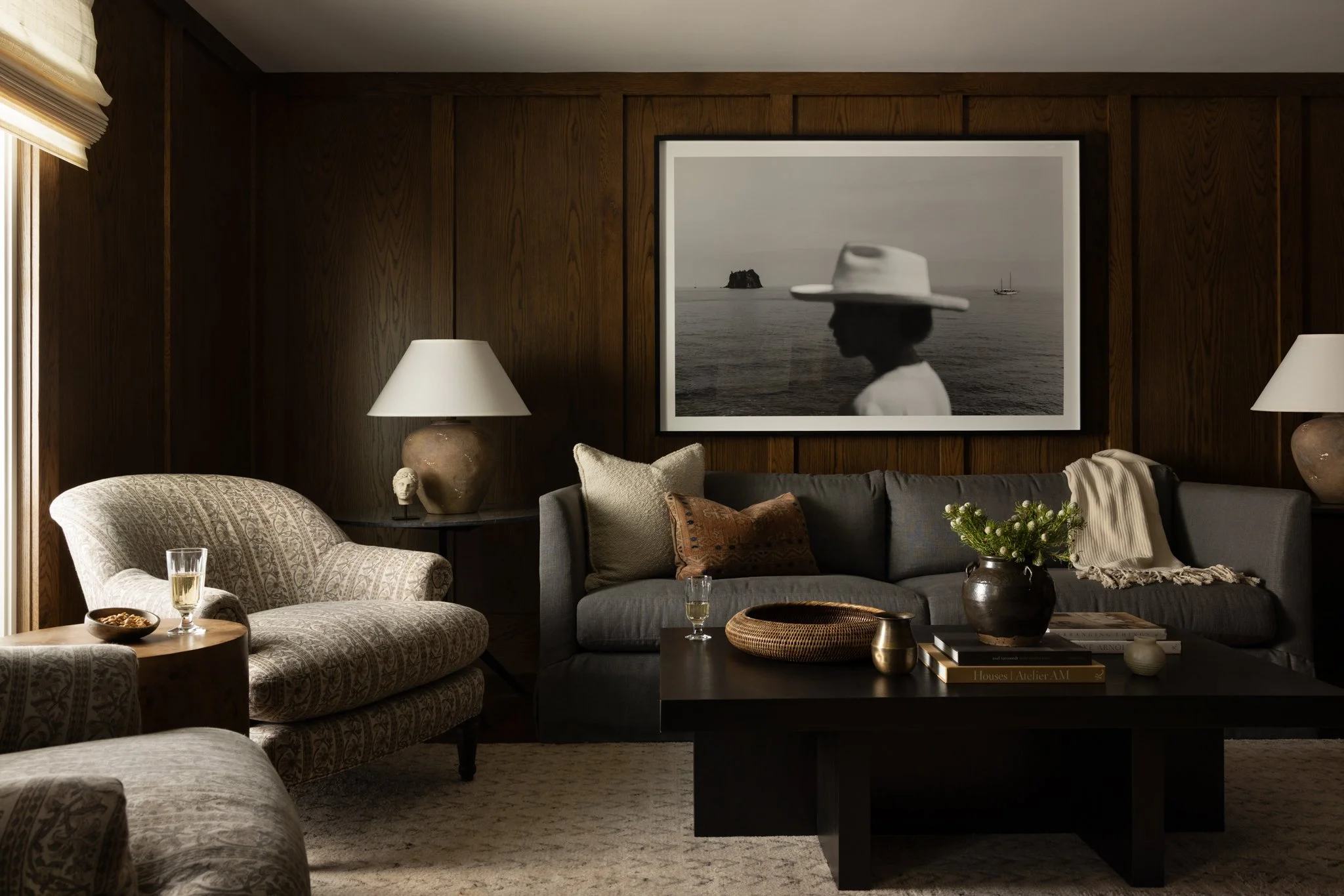 A living room with dark wood-paneled walls, a gray sofa with cushions, a patterned armchair, a coffee table with books, a vase with flowers, lamps, and a black-and-white framed photograph of a woman in a hat by the ocean.