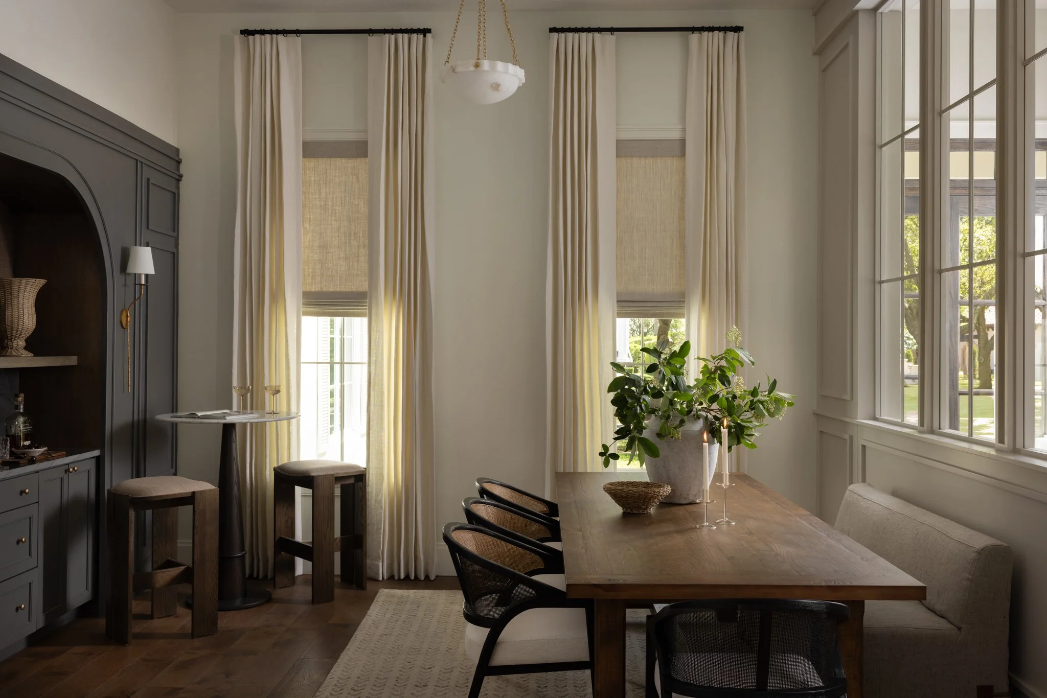 A bright dining room with a wooden table, black chairs, and beige wall bench. A large potted plant with greenery in the center of the table, with candles next to it. Large windows with cream-colored curtains and shades, allowing natural light.