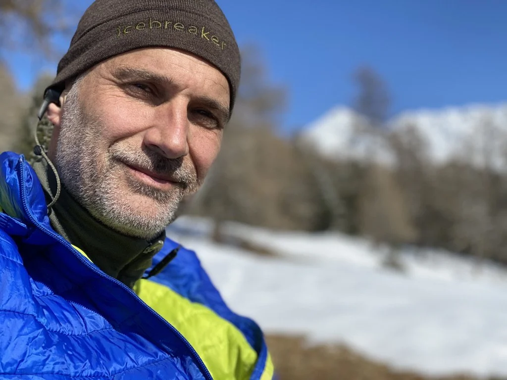 Marc souriant portant un bonnet gris et une veste de outdoor, avec un paysage enneigé en arrière-plan.