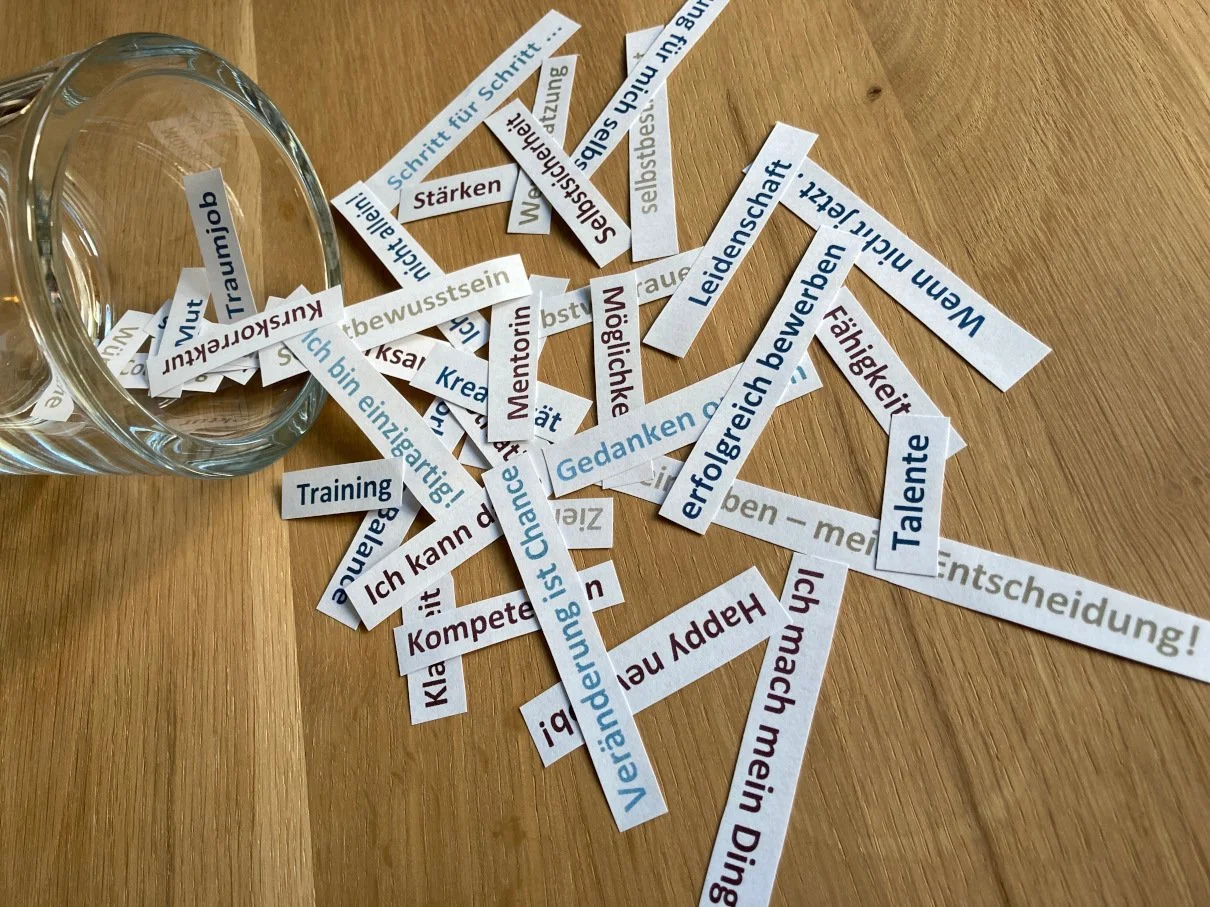 Zettel mit deutschen und englischen Worten und Phrasen, die aus einem Glas gefallen sind, auf einem Holztisch.