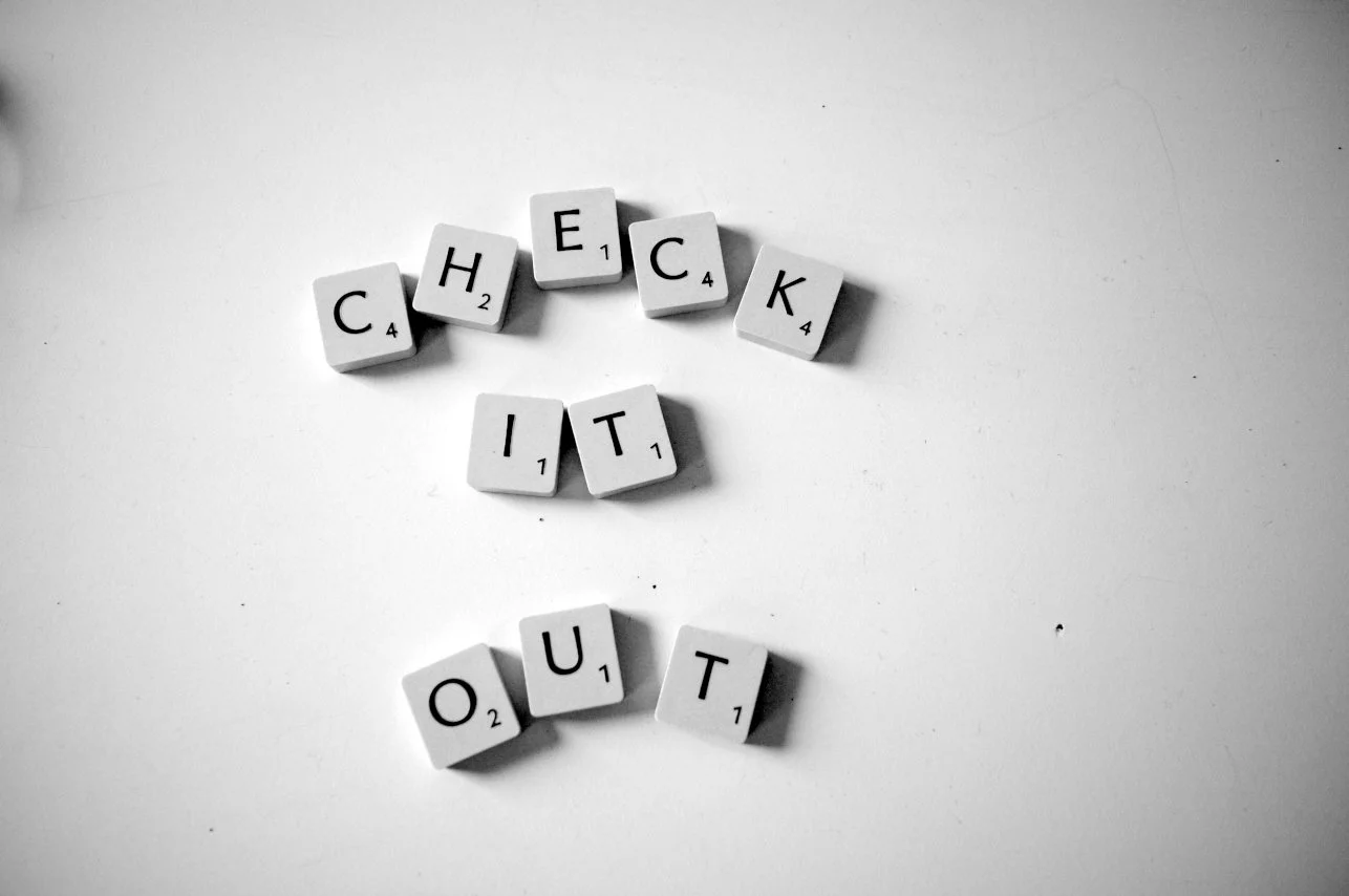 Schriftzug aus Scrabble-Steinen, der 'CHECK' und 'IT' sowie 'OUT' bildet, in Schwarz-Weiß.