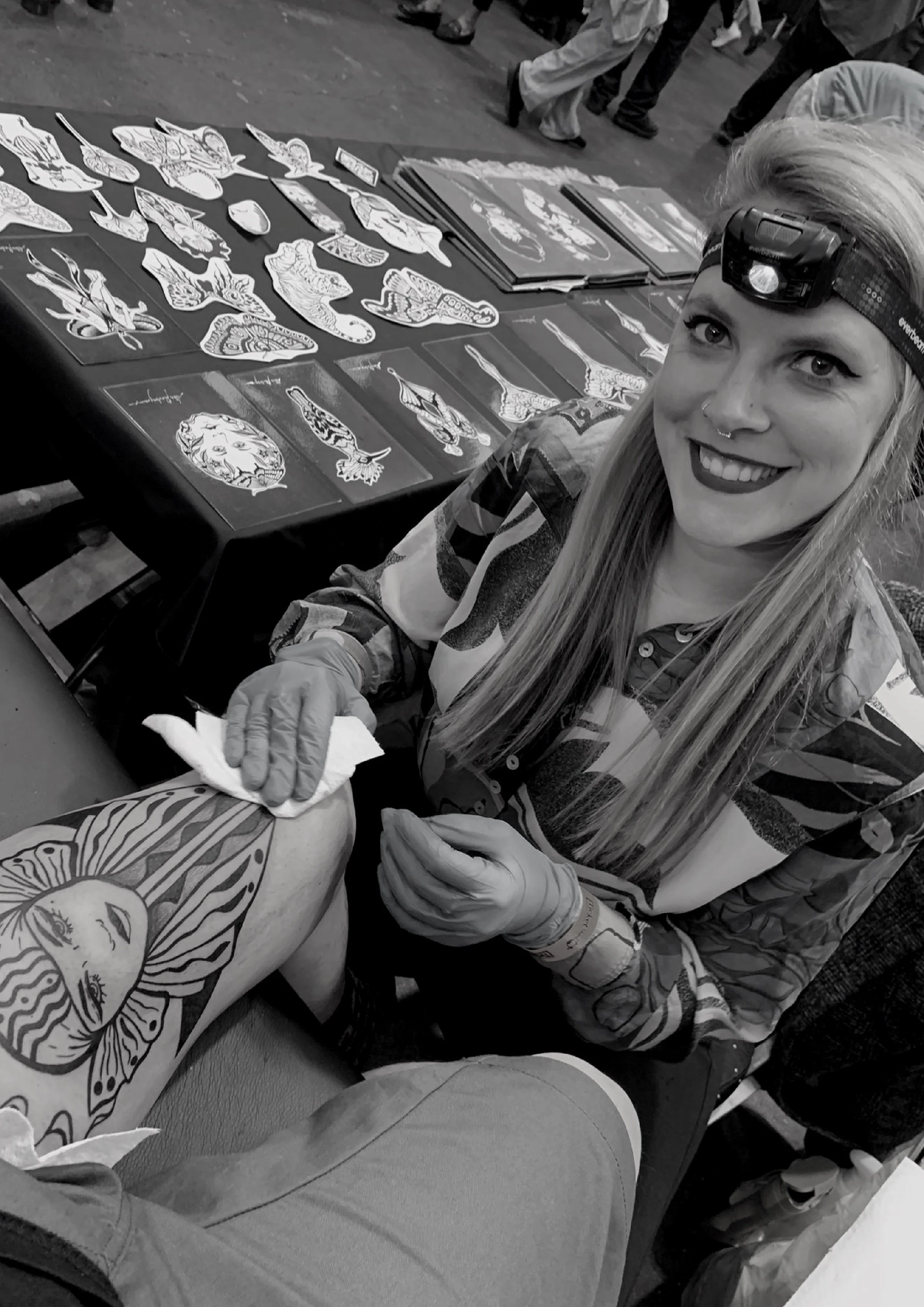 A woman with long hair, tattoos, and a nose piercing sitting at a table with tattoo flash sheets, wearing a headlamp and gloves, at a tattoo convention or event.