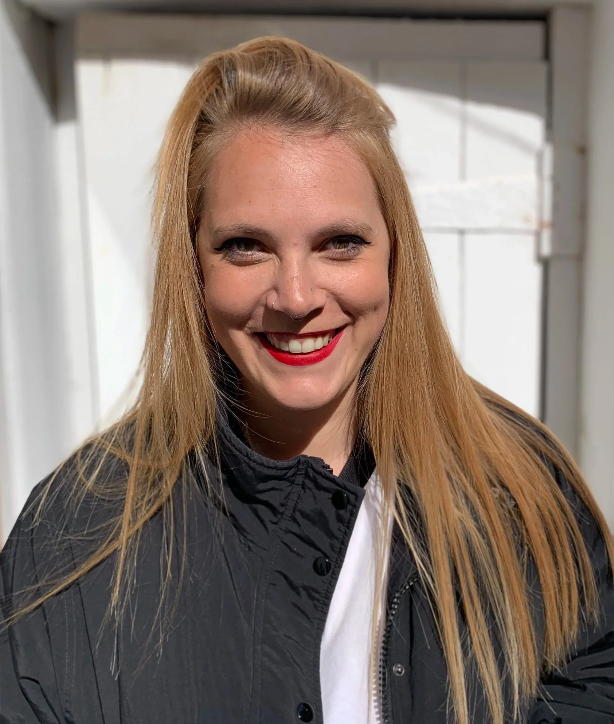 A woman with long blonde hair smiling, wearing red lipstick, a nose ring, a black quilted jacket, and a white shirt, standing outdoors in front of a white brick wall.