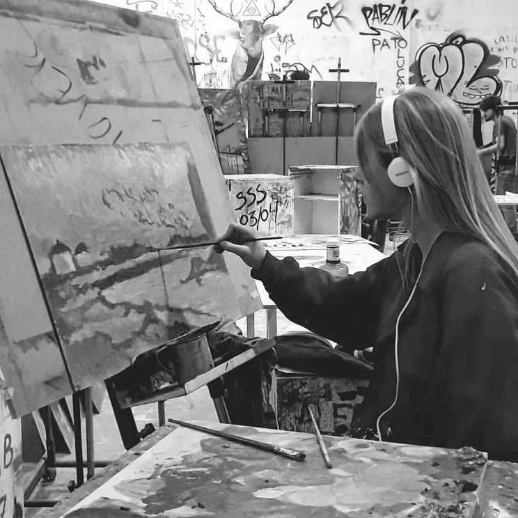 A woman with long hair wearing headphones, painting on a canvas in an art studio with graffiti on the walls.