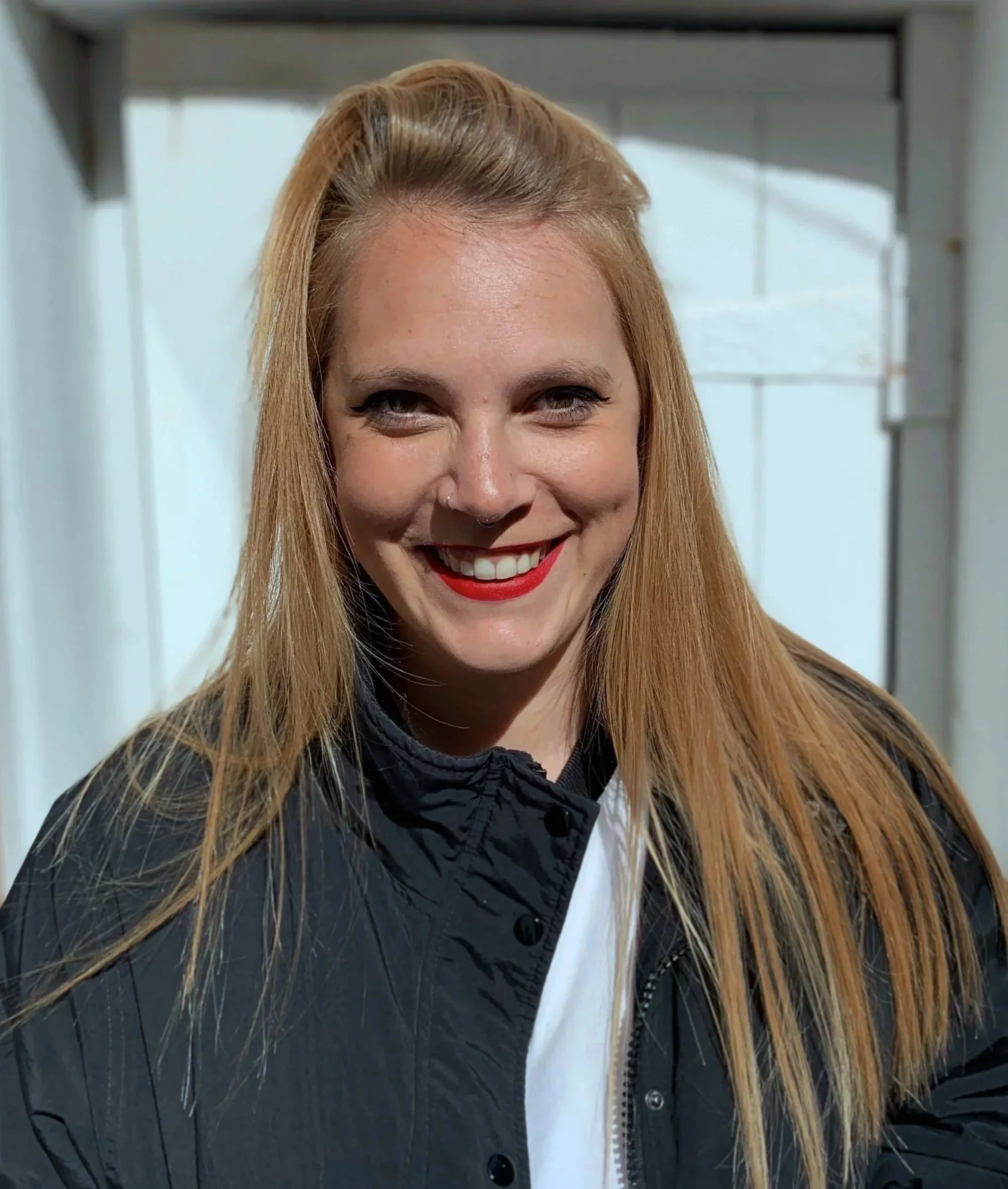 Close-up of a smiling woman with long blonde hair, wearing makeup with red lipstick, a black jacket, and a white shirt, standing in front of a blurred industrial background.