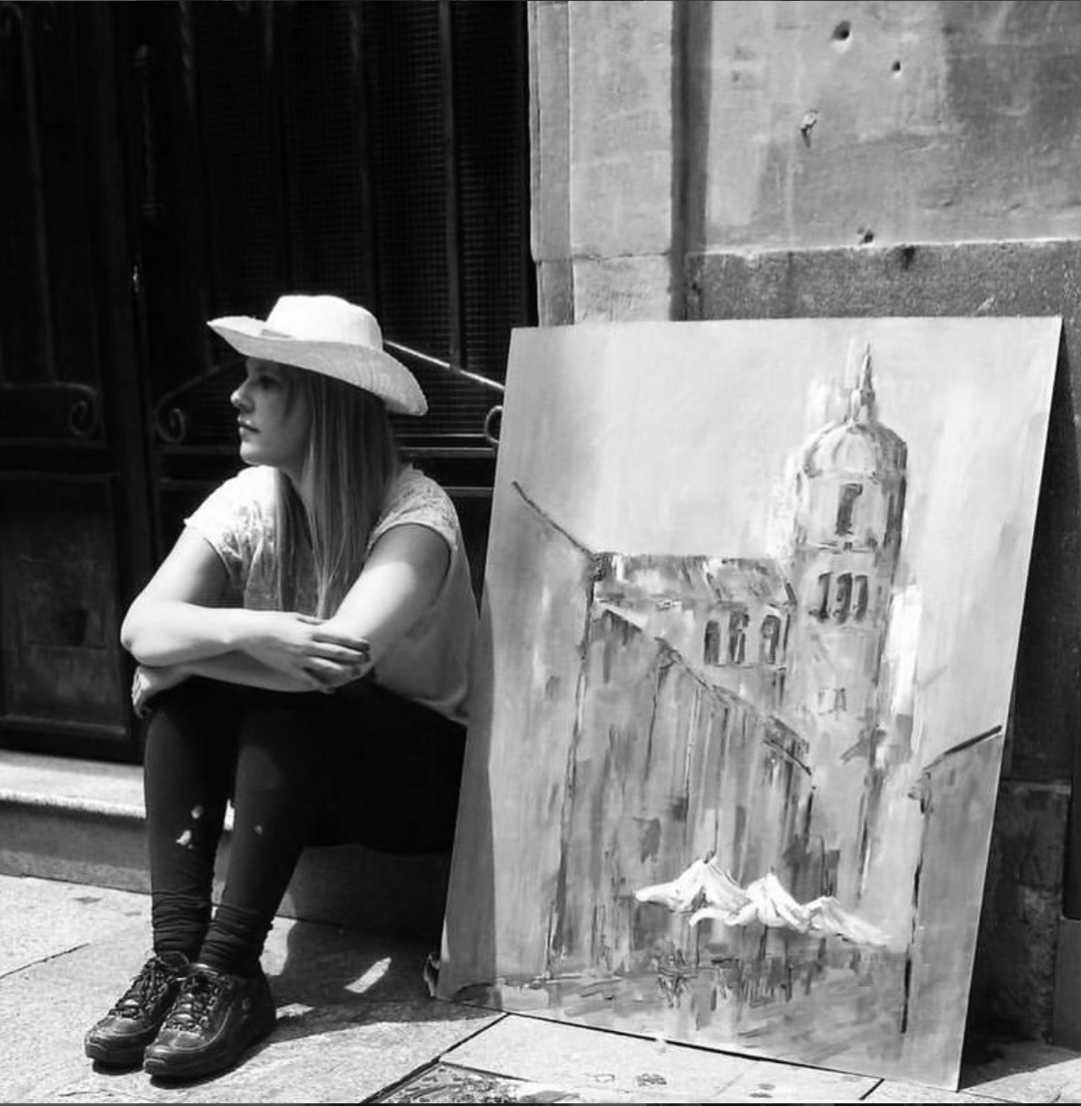 A woman wearing a wide-brimmed hat sitting on the ground next to an easel with a painting of a cityscape and a prominent clock tower.