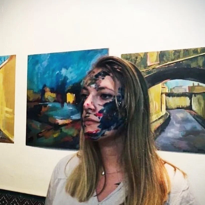Woman with face paint standing in front of three colorful landscape paintings in an art gallery.