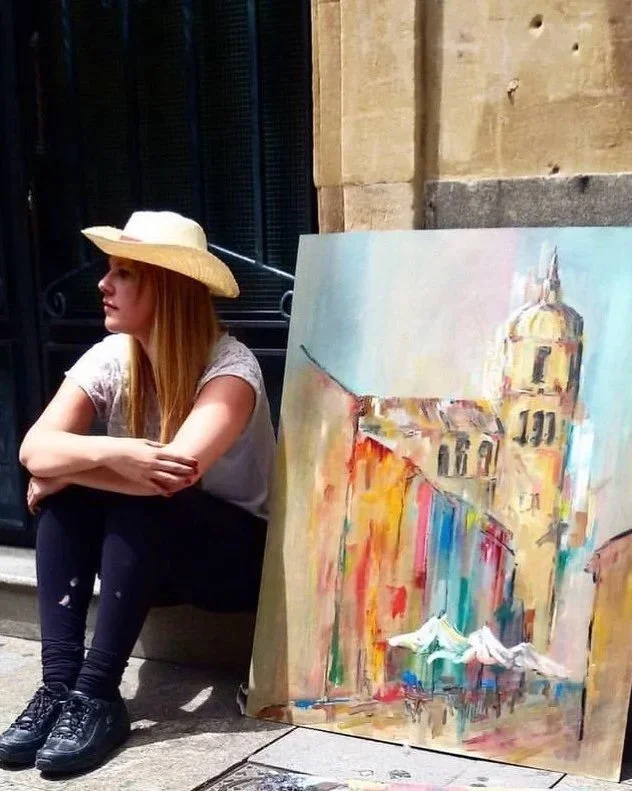 A woman with long red hair wearing a white hat, gray shirt, black pants, and black shoes sitting on the sidewalk near a colorful painting of a historic building with a tower and outdoor market stalls.