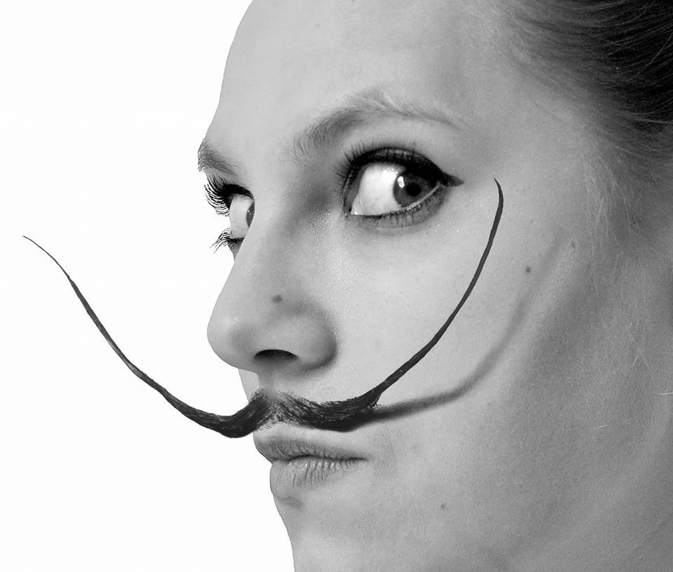 Close-up black-and-white photo of a woman with drawn mustache and beard, looking to her right with a serious expression.