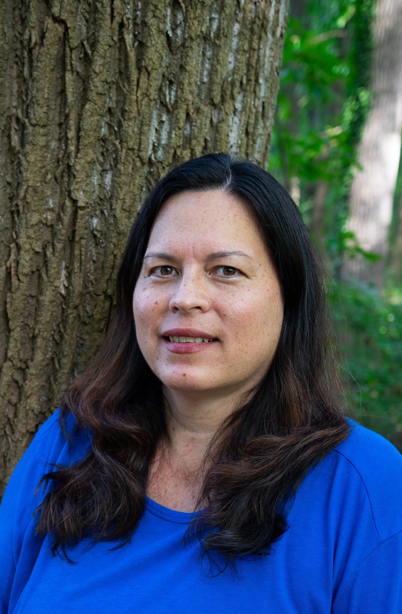 A woman with long dark hair and fair skin wearing a blue shirt, standing outdoors near a large tree with green foliage in the background.