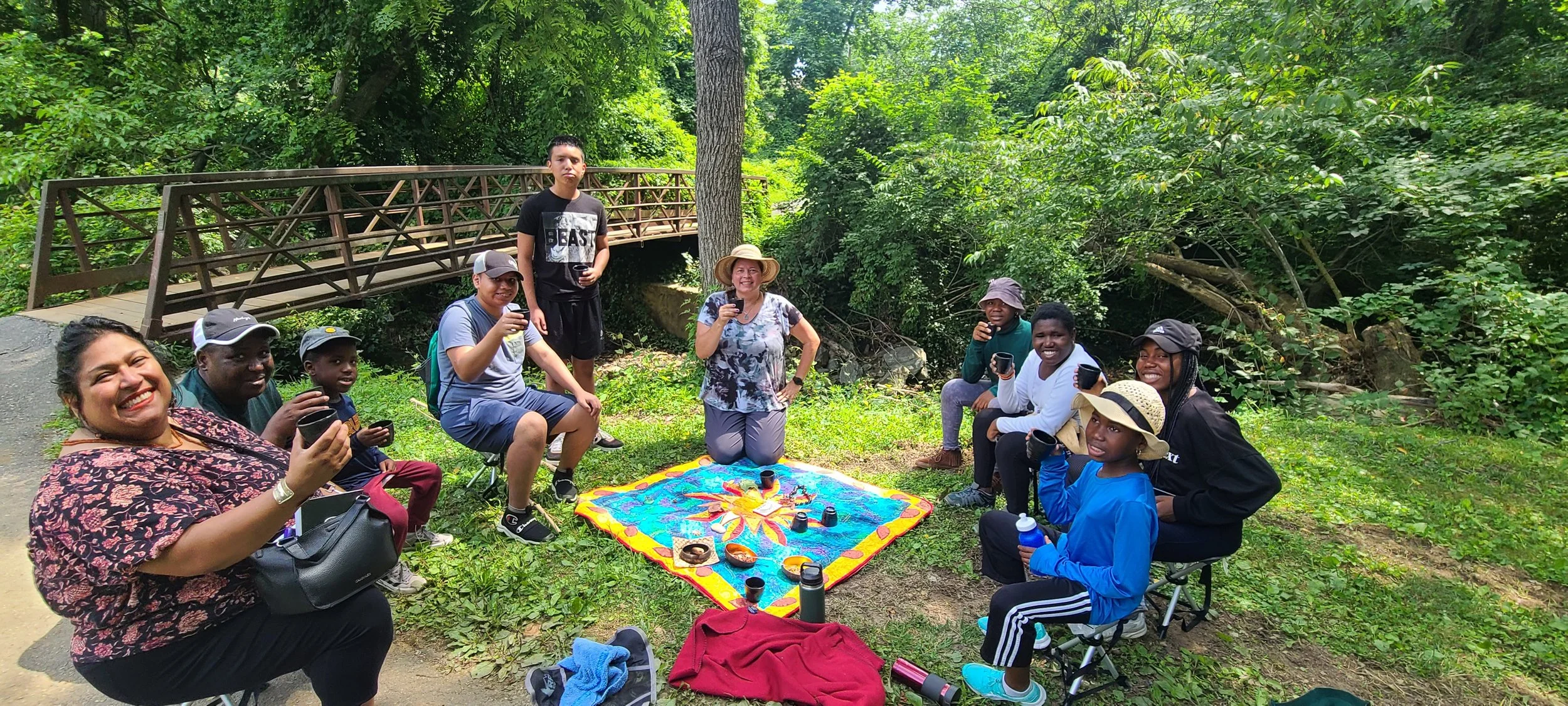 Group of people outdoors in a forest, sitting and standing around a colorful blanket with food and drinks, smiling and enjoying each other's company.