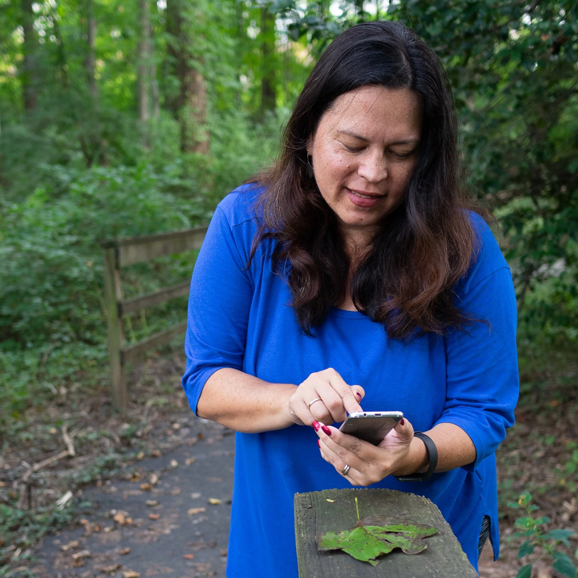 Woman with dark hair wearing a blue jacket and red nail polish, standing outside in a green wooded area, looking at her smartphone while her left hand rests on a weathered wooden post with leaves, and a trail with fallen leaves is visible in the background.