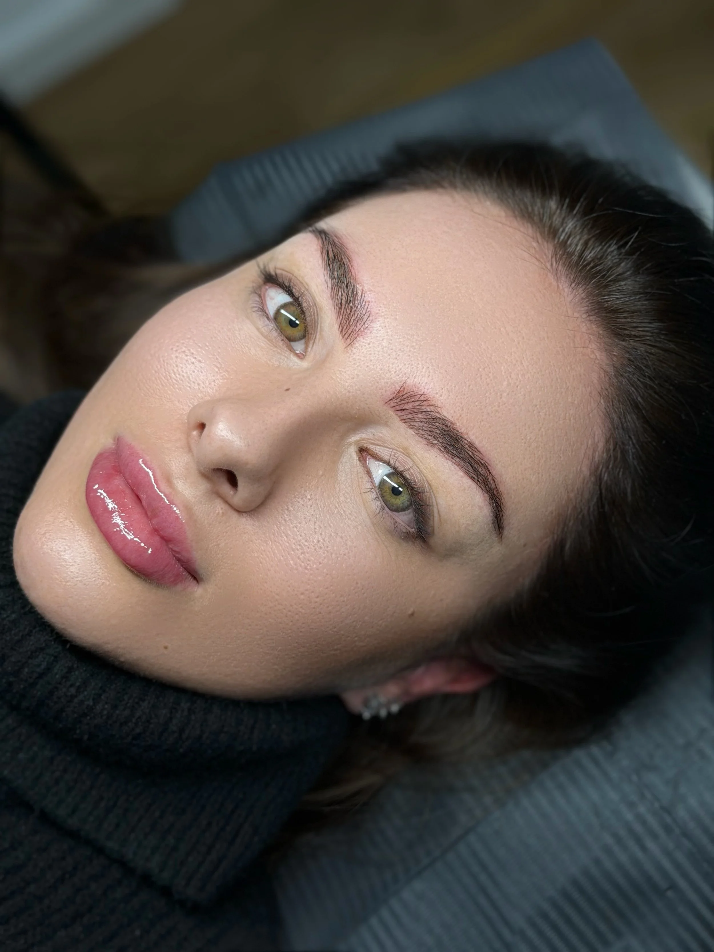 Nahaufnahme eines jungen Frauengesichts mit blondierten Lippen, klarer Haut, dunklen Haaren, und braunen Augen, liegend auf einer grauen Unterlage, getragen von einer Person in schwarzem Pullover.