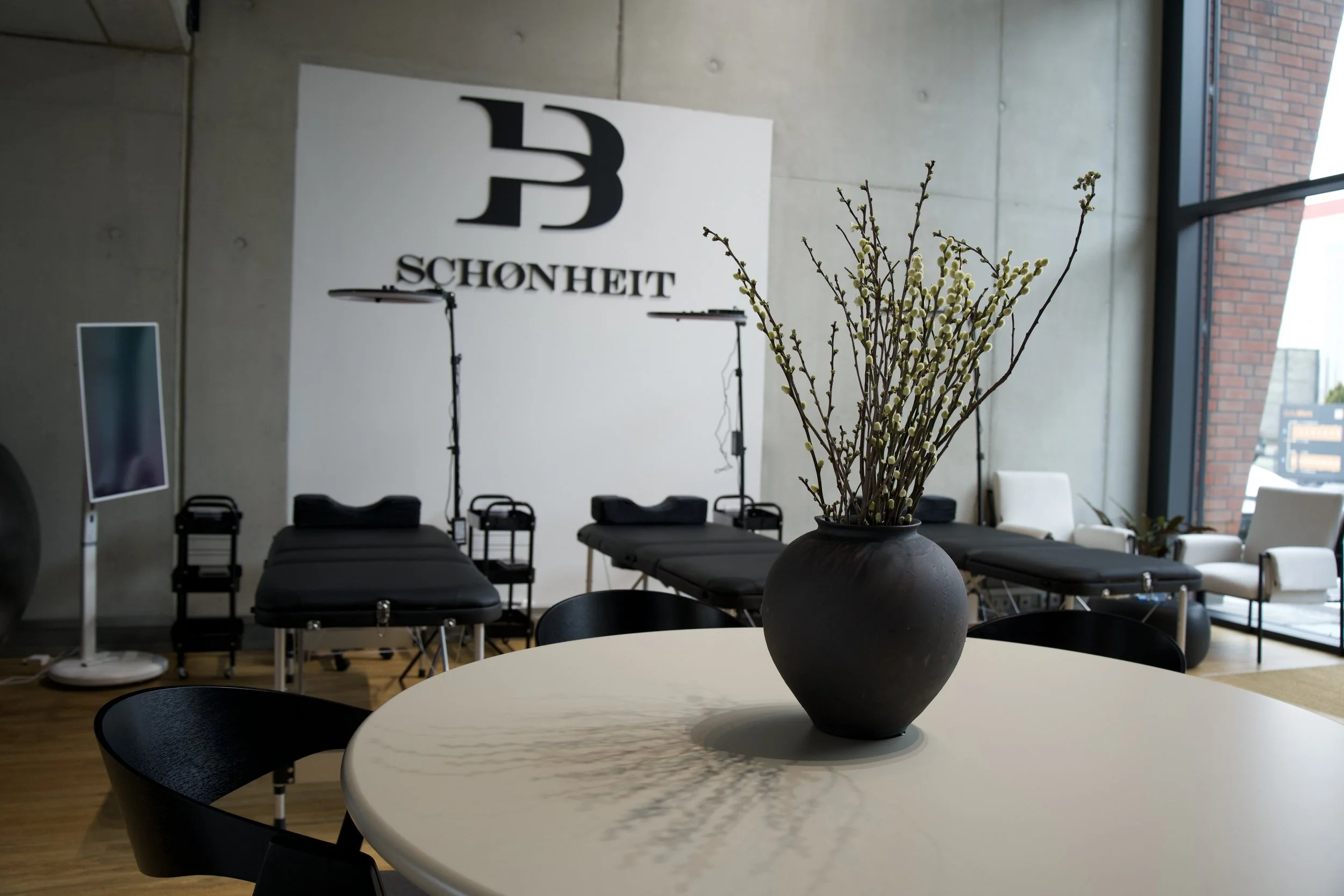 Ein modernes Raum mit einem runden Tisch, auf dem eine schwarze Vase mit Zweigen steht. Im Hintergrund sind Liegen und ein Logo mit der Aufschrift 'SCHONHEIT' an der Wand, sowie großen Fenstern, die Tageslicht in den Raum lassen.