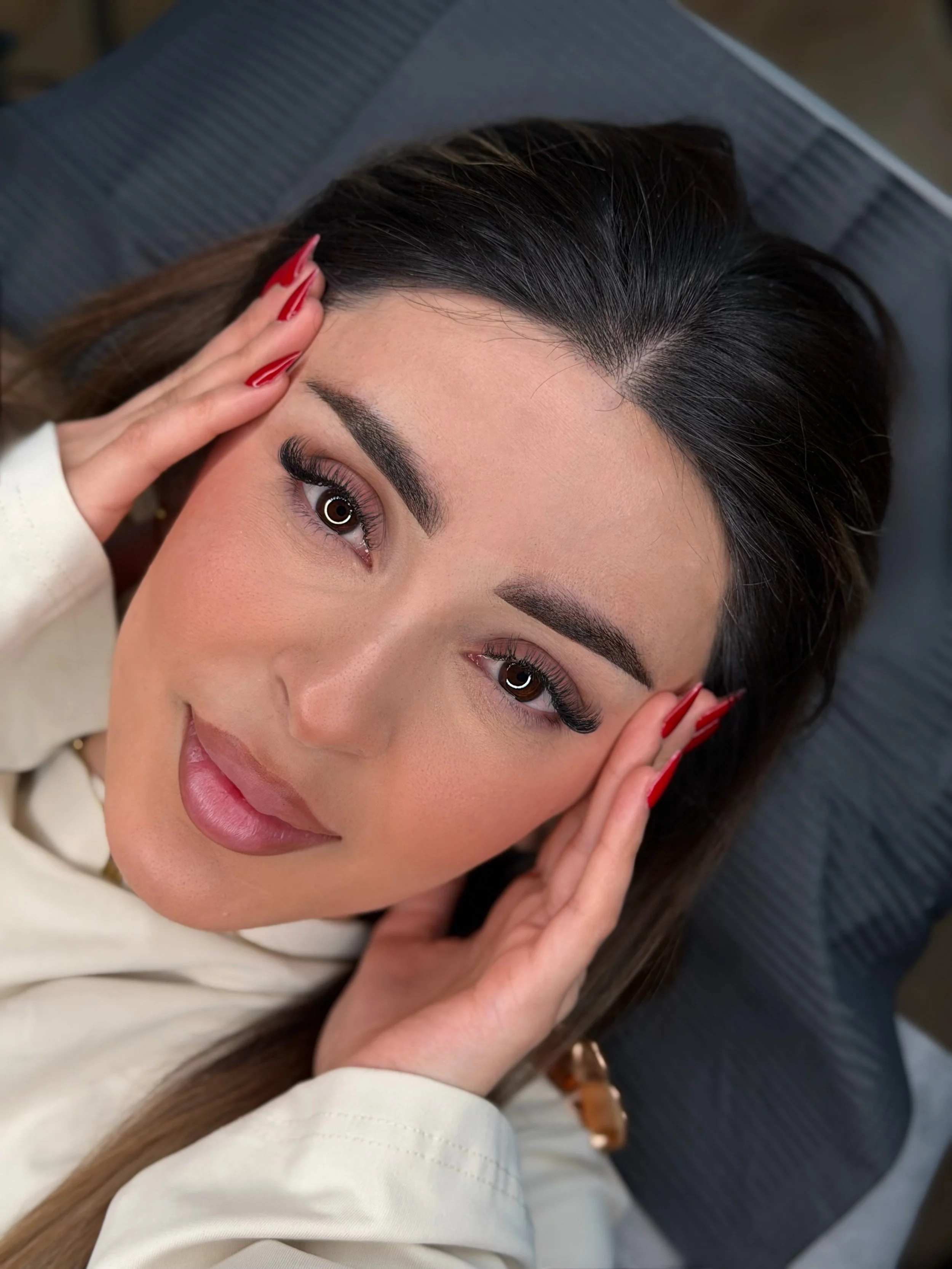 Nahaufnahme einer lächelnden Frau mit dunklem Haar, braunen Augen, gepflegten Nägeln und Make-up, liegend auf einem grauen Kissen.