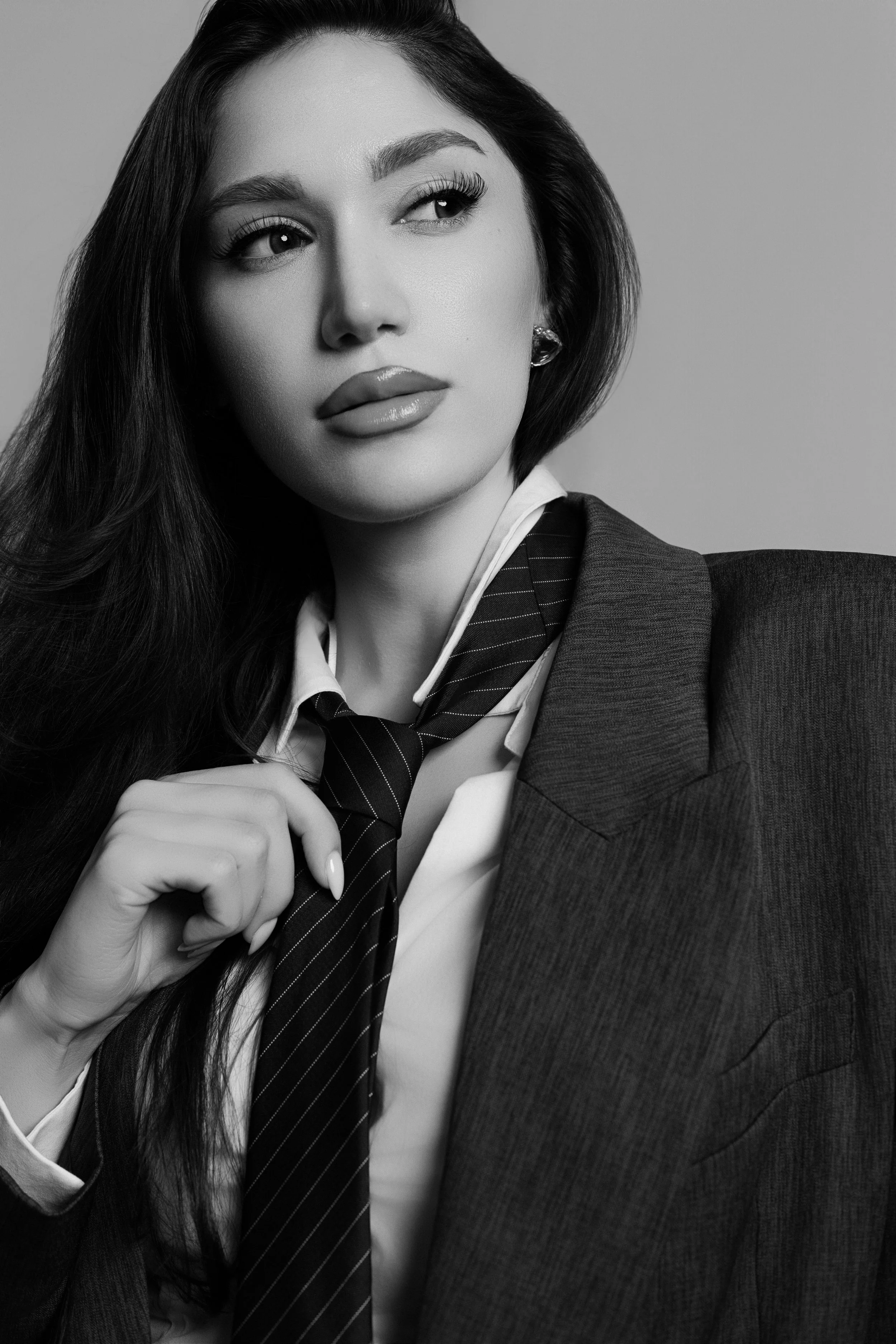 Schwarz-weiß Fotografie eines jungen, eleganten Mädchens mit langen Haaren, die einen schwarzen Anzug und Krawatte trägt, und auf einem Stuhl sitzt.