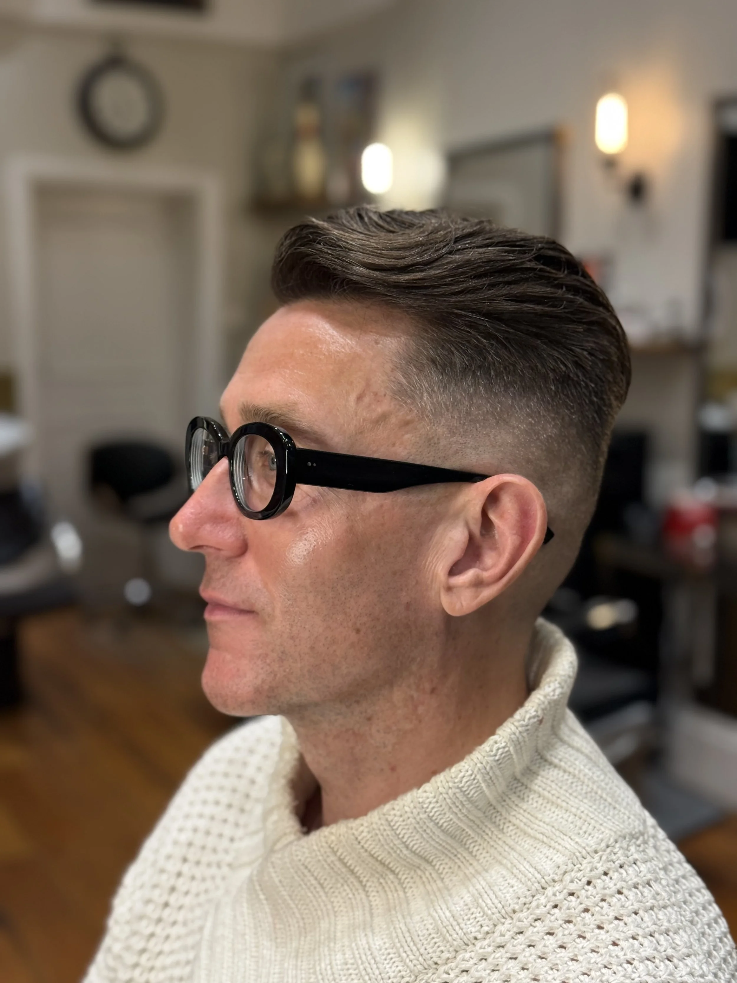 Close-up of a man's side profile with styled brown hair, dark glasses, and wearing a cream-colored knitted sweater, in a cozy indoor setting.