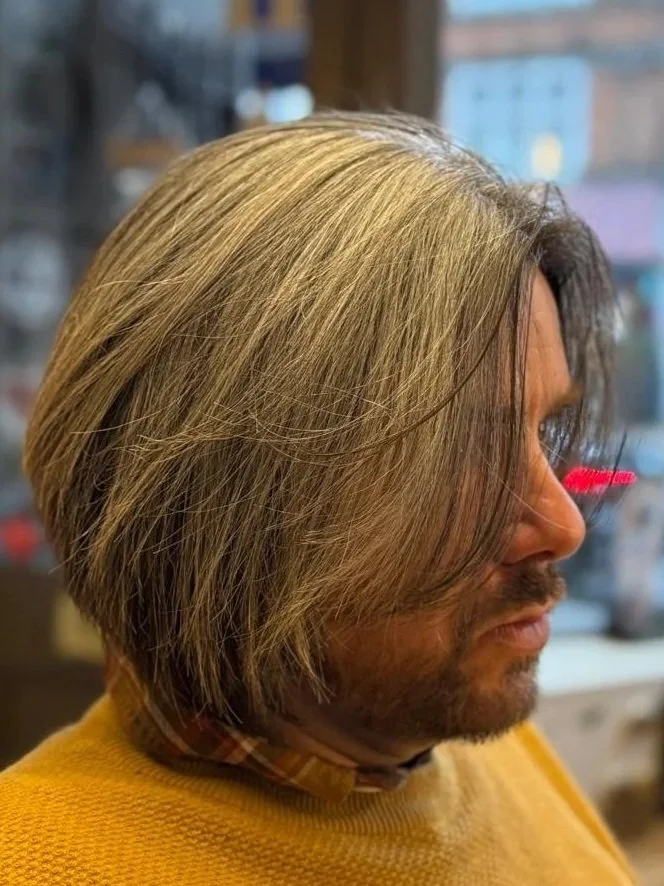 A man with long, layered, blonde hair, wearing a yellow sweater and reading glasses, looking to the right, in an indoor setting.