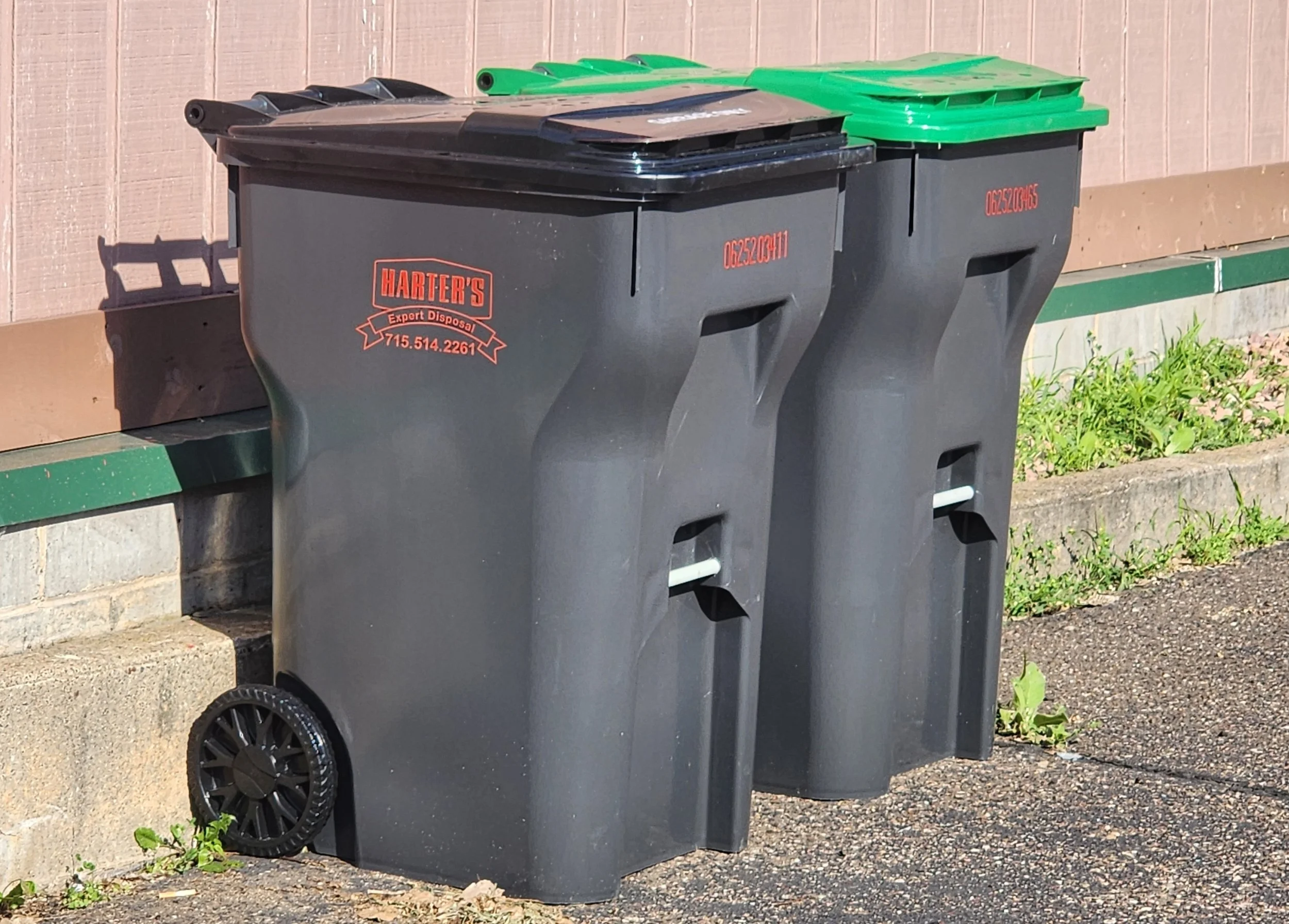 Two trash and recycling bins, one black and one green, are placed side by side outdoors against a pinkish wall. The black bin has red lettering with a business name and phone number.