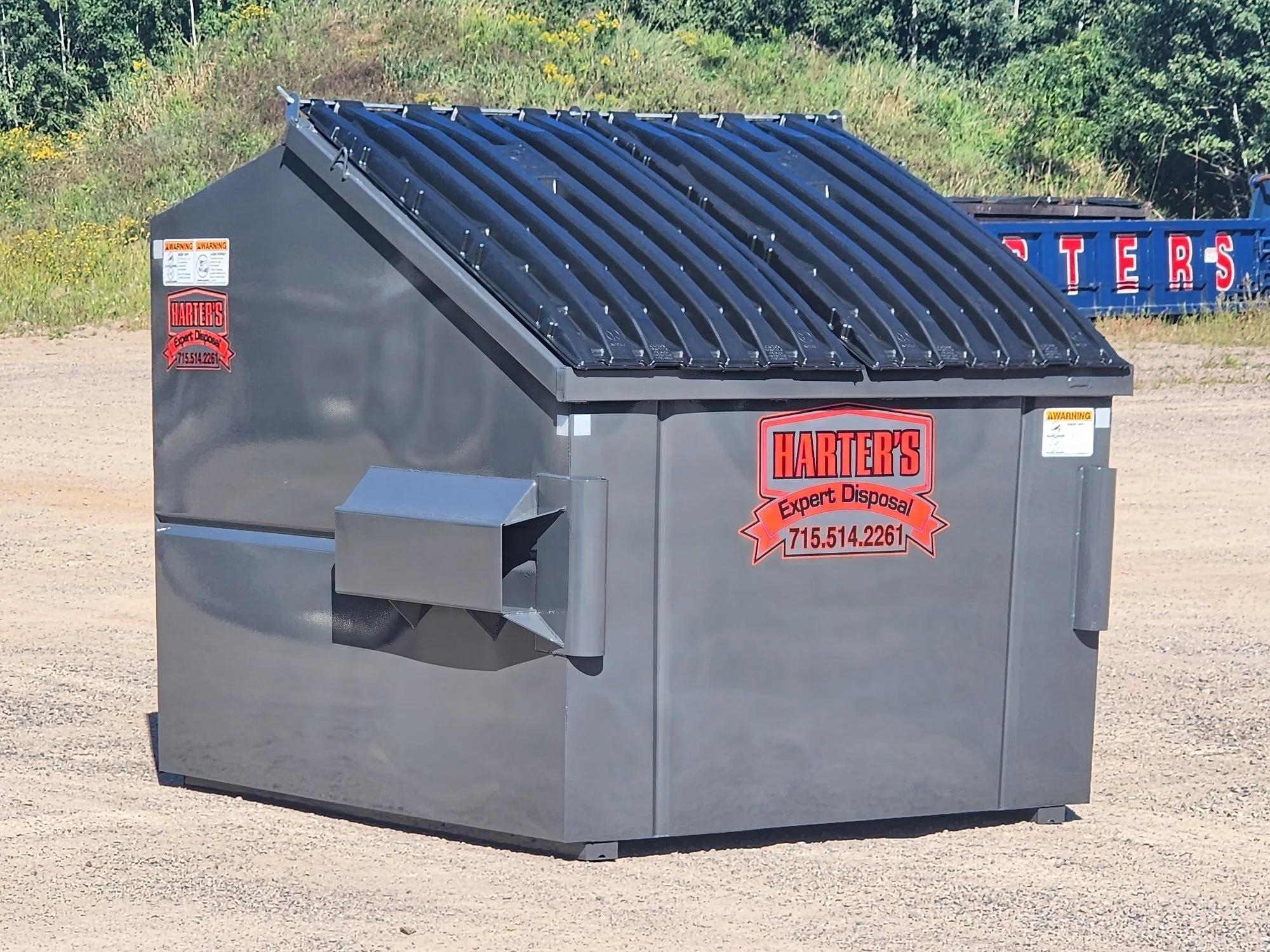 A gray metal dumpster with a black lid on a gravel lot, with warning labels and a company sticker, and a blue truck in the background with red lettering.