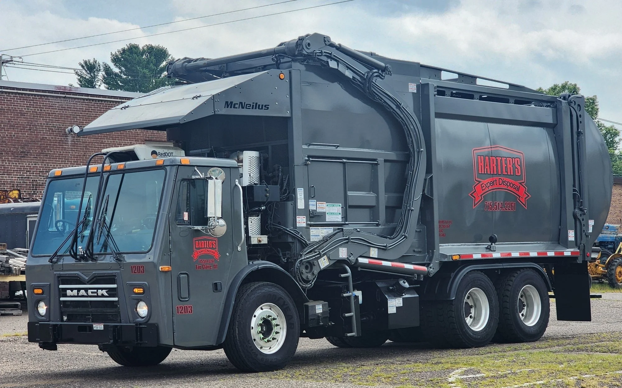 A gray garbage truck with the logo "Harter's Expert Disposal" on the side, parked outdoors near a brick building and trees under a cloudy sky.