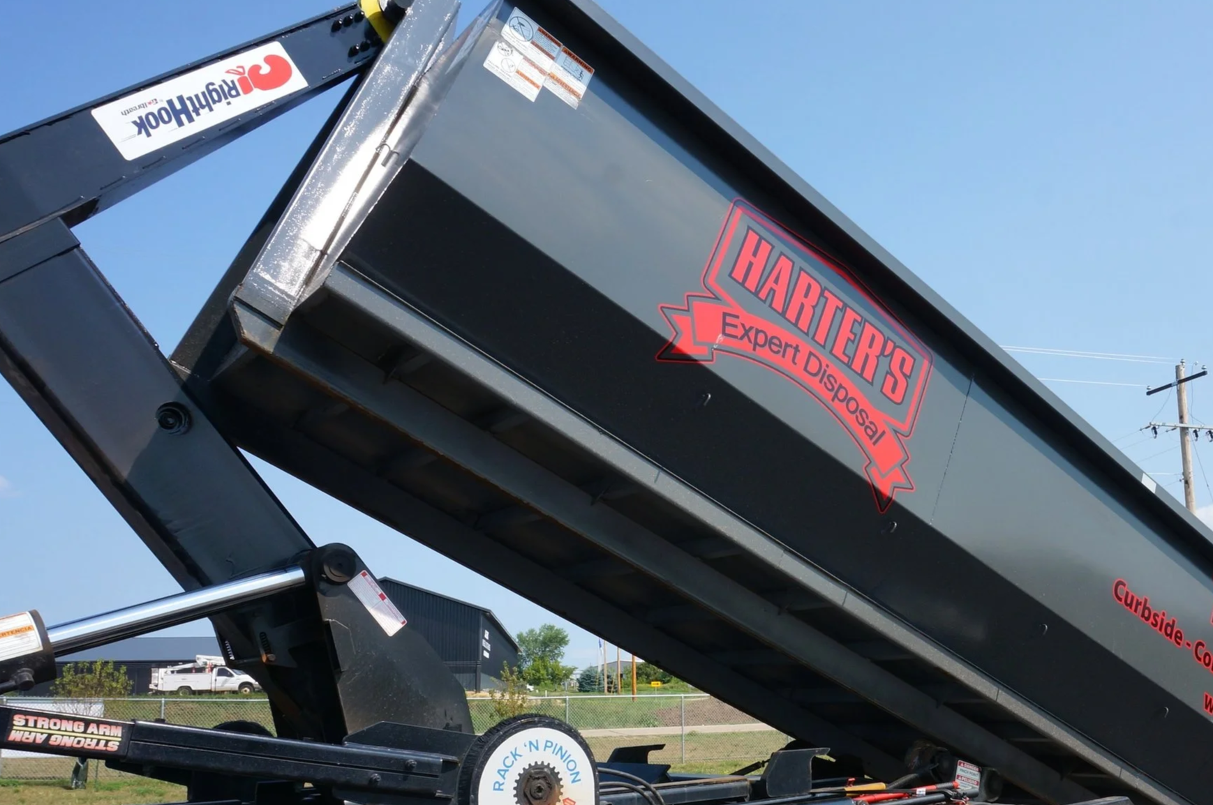Close-up of a black trash or recycling truck with red and black logo reading 'HARTIER'S Expert Disposal'. The truck is tilted upward with clear blue sky in background. Visible hydraulic mechanism and safety warnings.